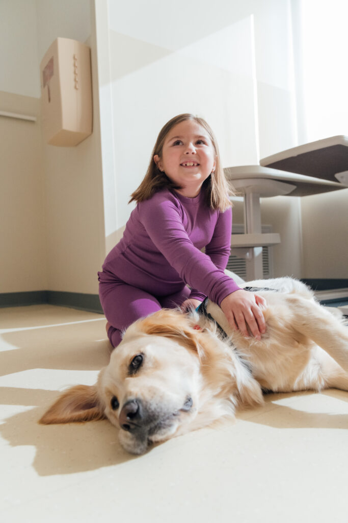 A young girl playing on the floor with one of Dayton Children’s Canine Co-pilots, laughing and smiling.
