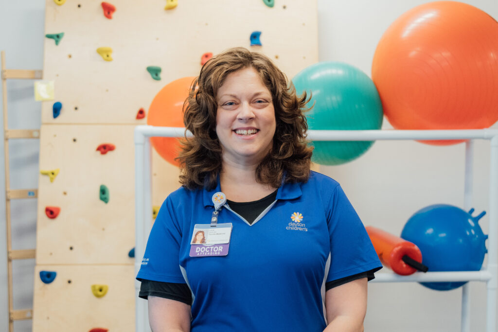 A female healthcare professional, smiling, stands in a brightly lit gym, wearing a blue polo shirt and a "Doctor" badge, with large exercise balls and a climbing wall behind her.
