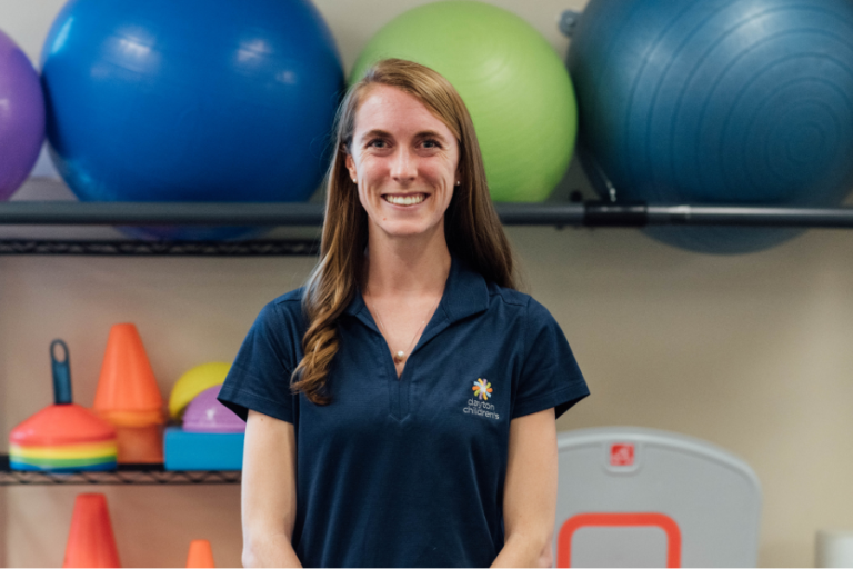 A smiling woman with long brown hair, wearing a dark blue polo shirt and a delicate necklace, stands in a brightly lit gym with exercise balls behind her.