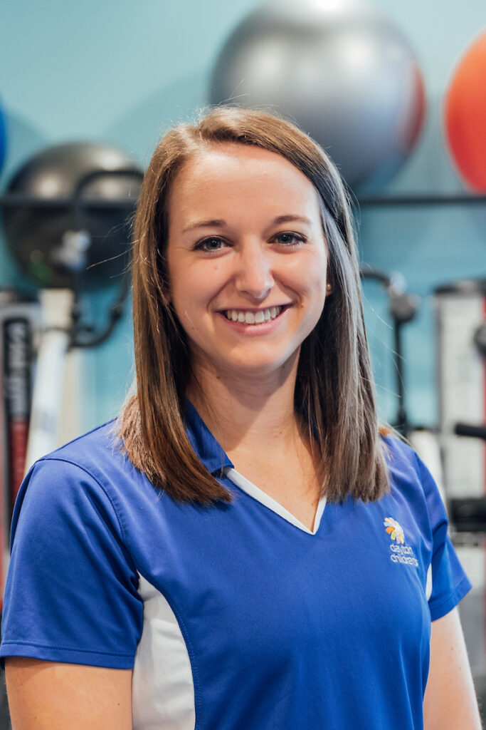 A professional headshot of a smiling woman with straight shoulder-length brown hair, wearing a blue and white athletic polo shirt in a gym setting.