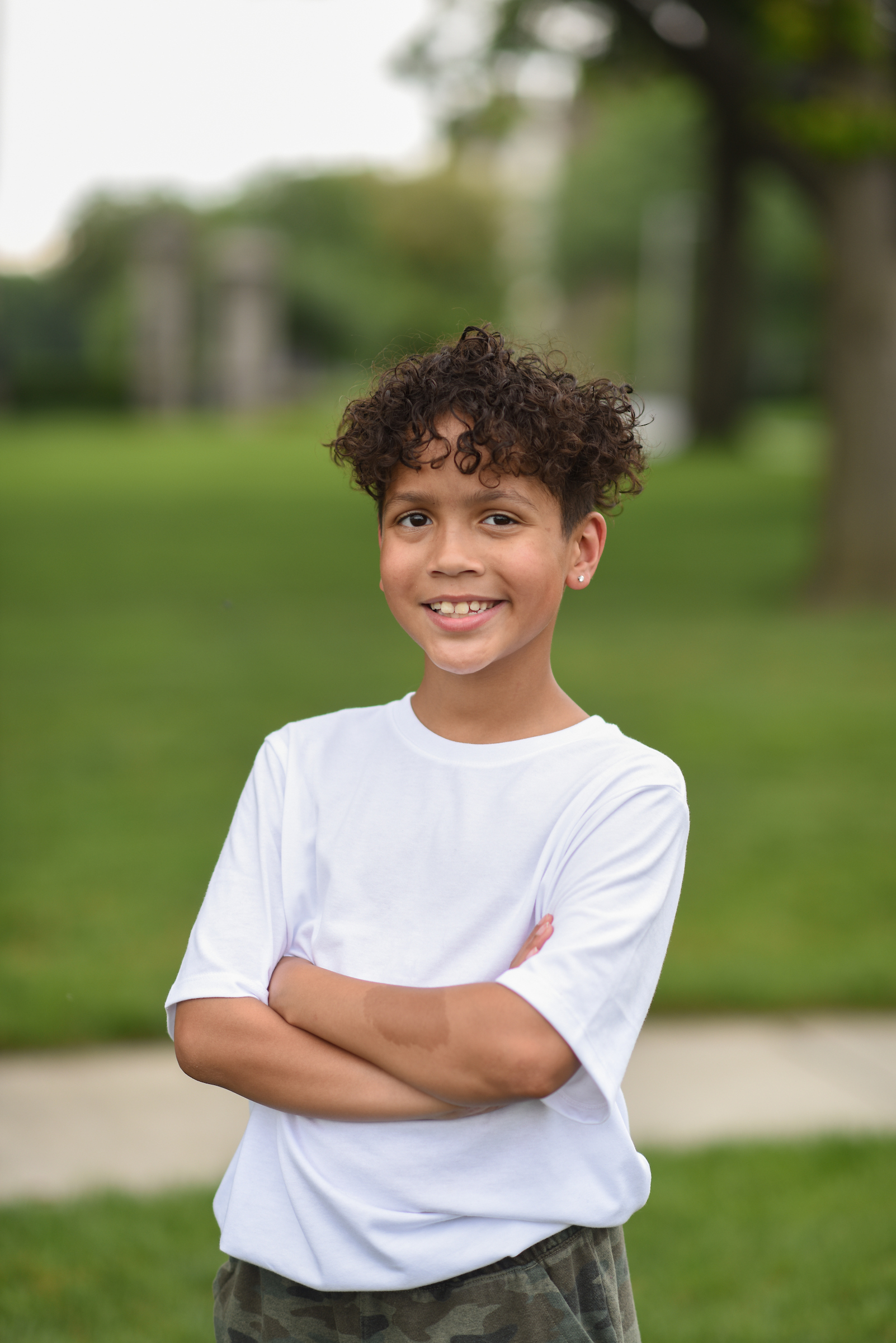 Smiling child standing outdoors with arms crossed, wearing a white shirt, representing a positive outcome after pediatric therapy.

