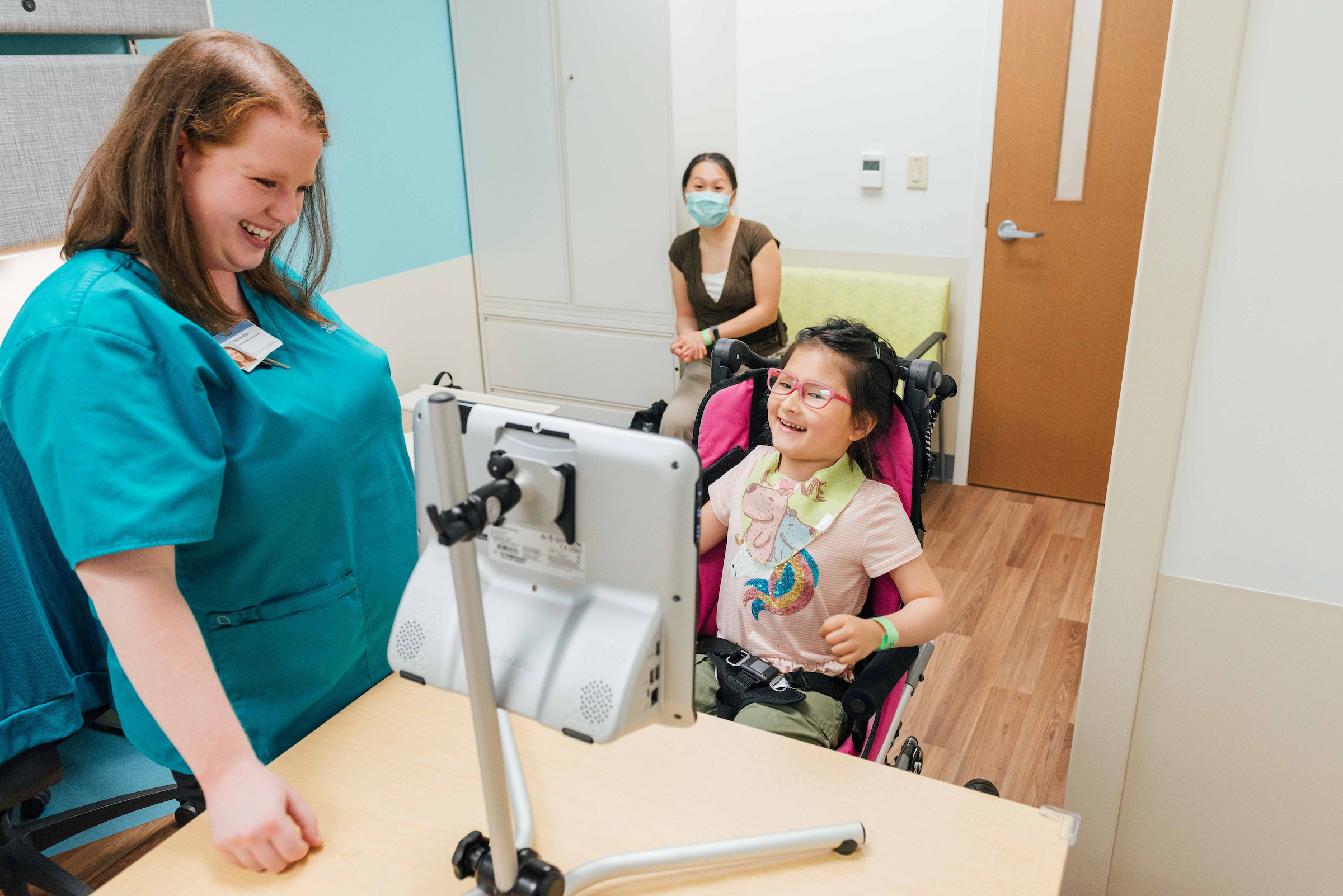 Pediatric therapist working with a young girl in a wheelchair using a communication device during a therapy session at a children’s hospital, with the child’s caregiver observing.
