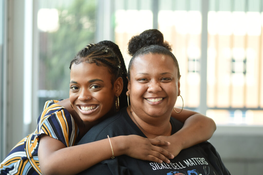 A teen sickle cell patient wraps her arms around her mom from behind, smiling at the camera.