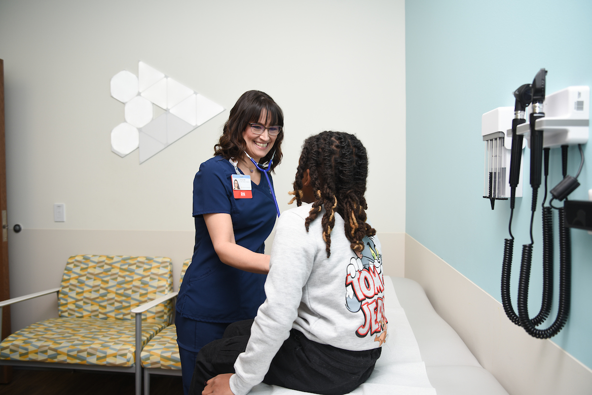A nurse in blue scrubs smiles while checking a child sitting on an exam table in a medical office.
