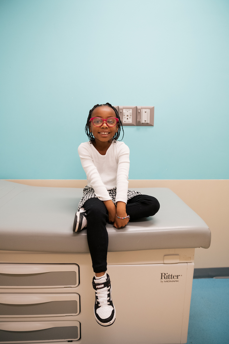 A young girl with pink glasses smiles while sitting on an exam table in a medical office.

