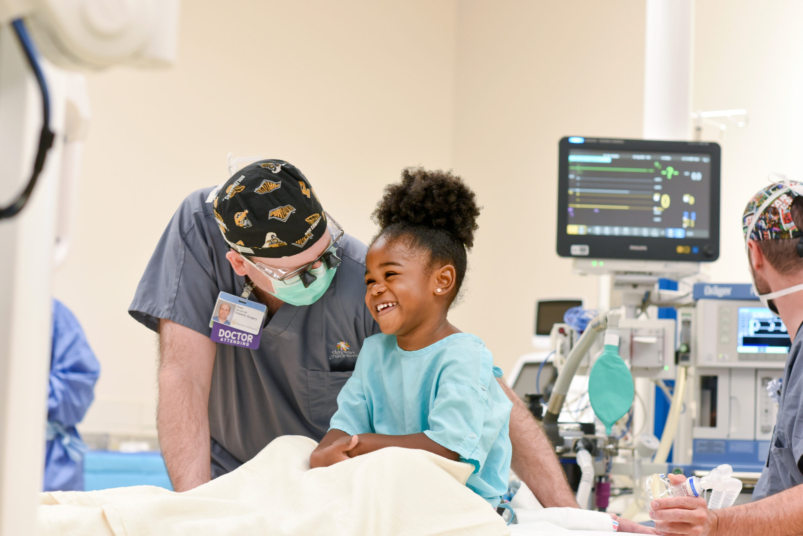 A doctor in scrubs and a mask smiles at a laughing young girl in a blue hospital gown on an operating table.
