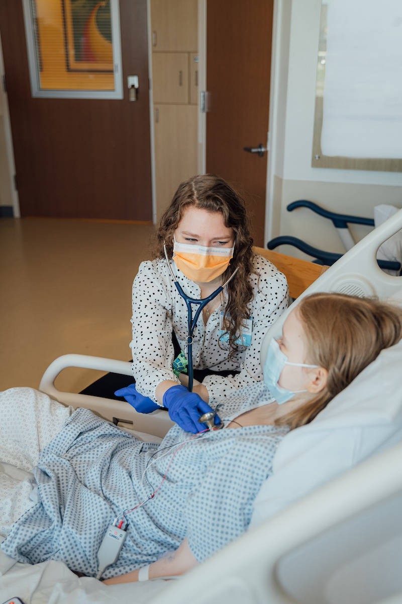 A masked doctor in a polka dot shirt uses a stethoscope to examine a masked patient in a hospital gown.