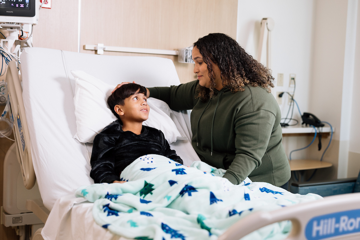 A caring woman with curly hair gently touches a young boy's head in a hospital bed, as he looks up at her.
