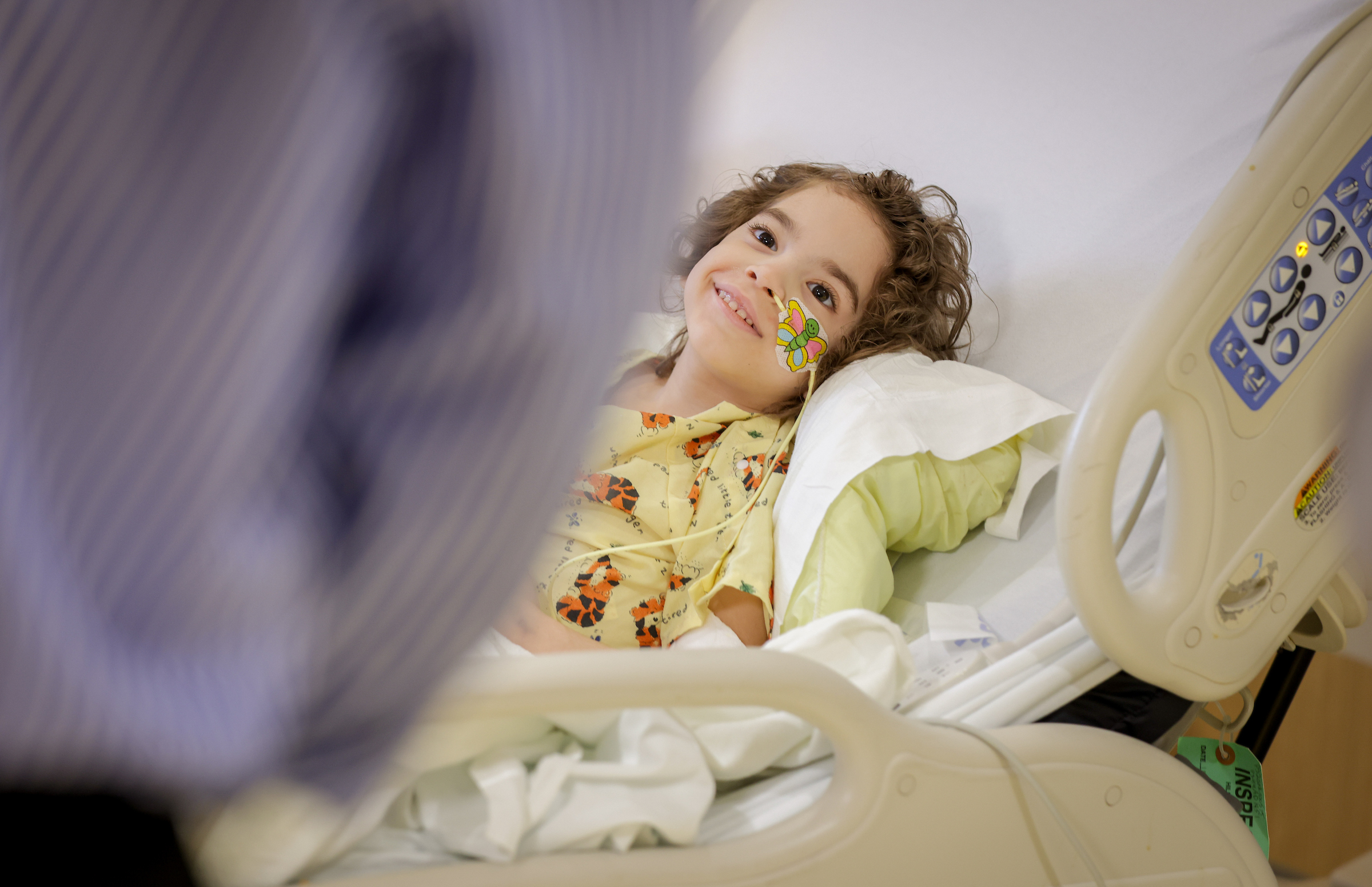 A smiling young girl with curly hair and a feeding tube lies in a hospital bed, looking towards a blurred person.
