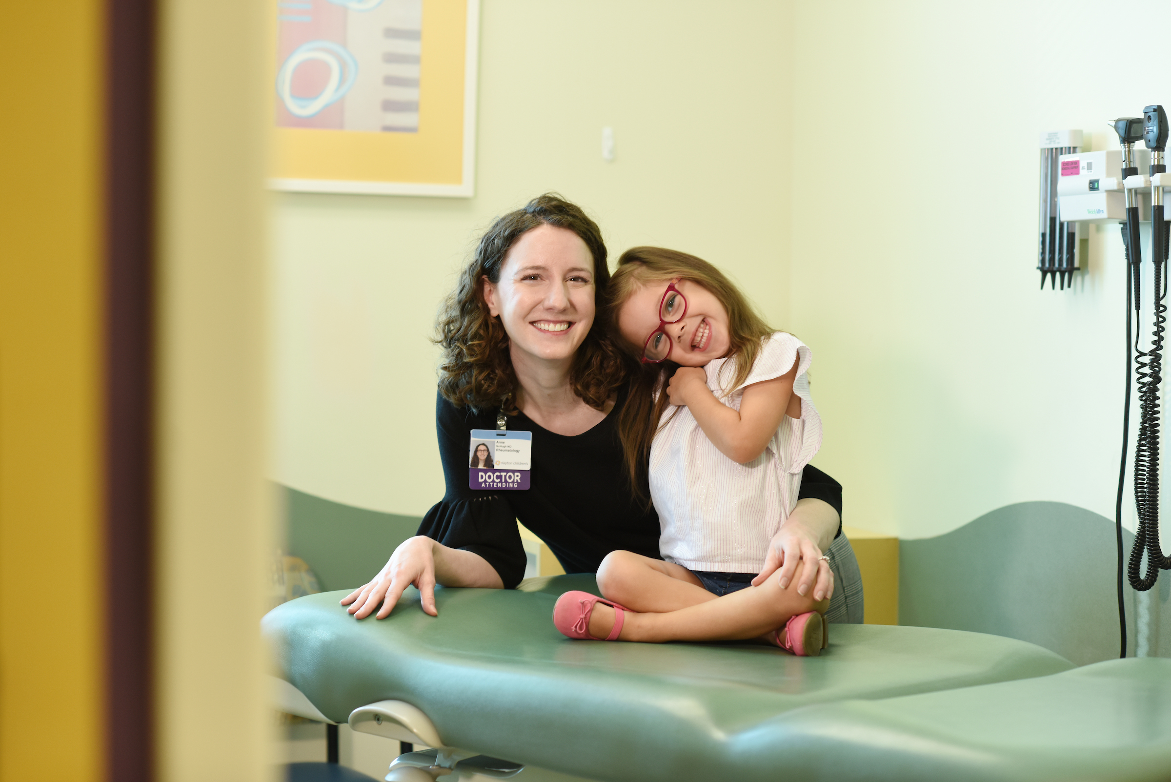 A smiling female doctor with curly hair and a "Doctor Attending" badge poses with a happy young girl in pink glasses, who leans on her playfully.

