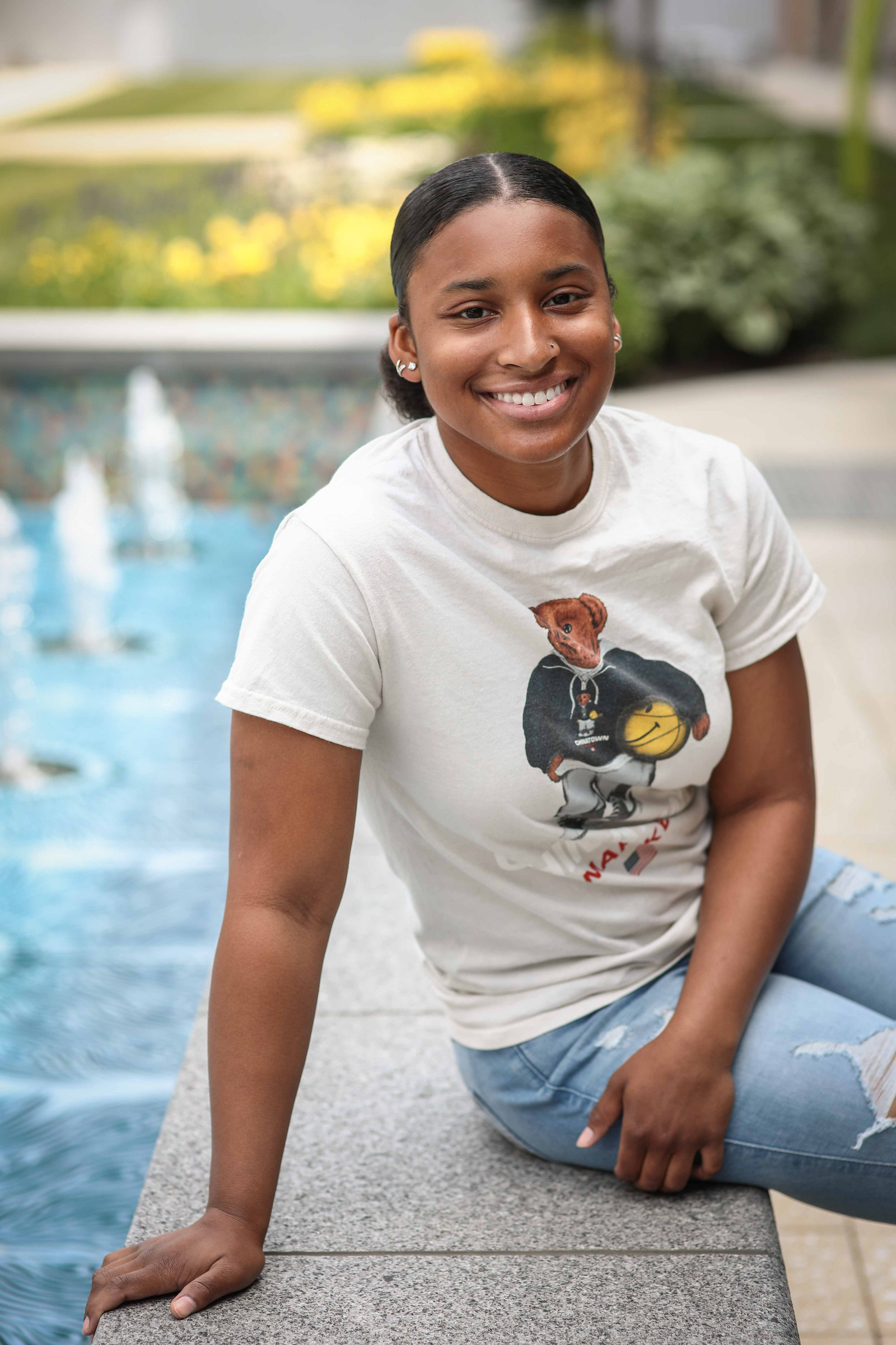 A young woman with dark hair pulled back, wearing a white t-shirt with a teddy bear graphic and light blue jeans, sits smiling by a fountain.

