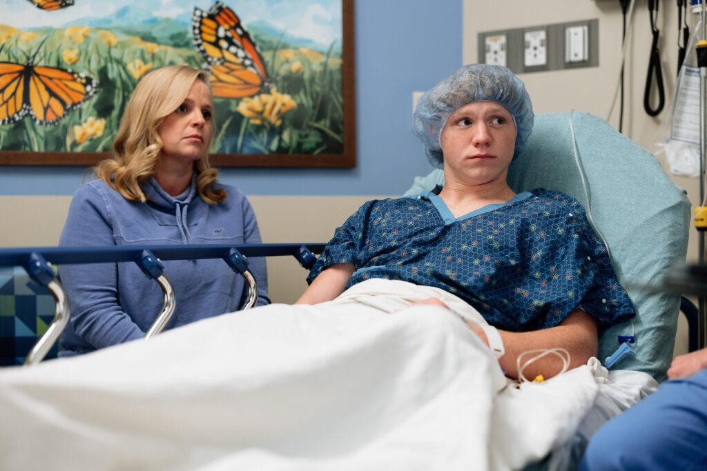 An anxious-looking patient in a hospital gown and hairnet sits in bed while a woman sits supportive next to them.
