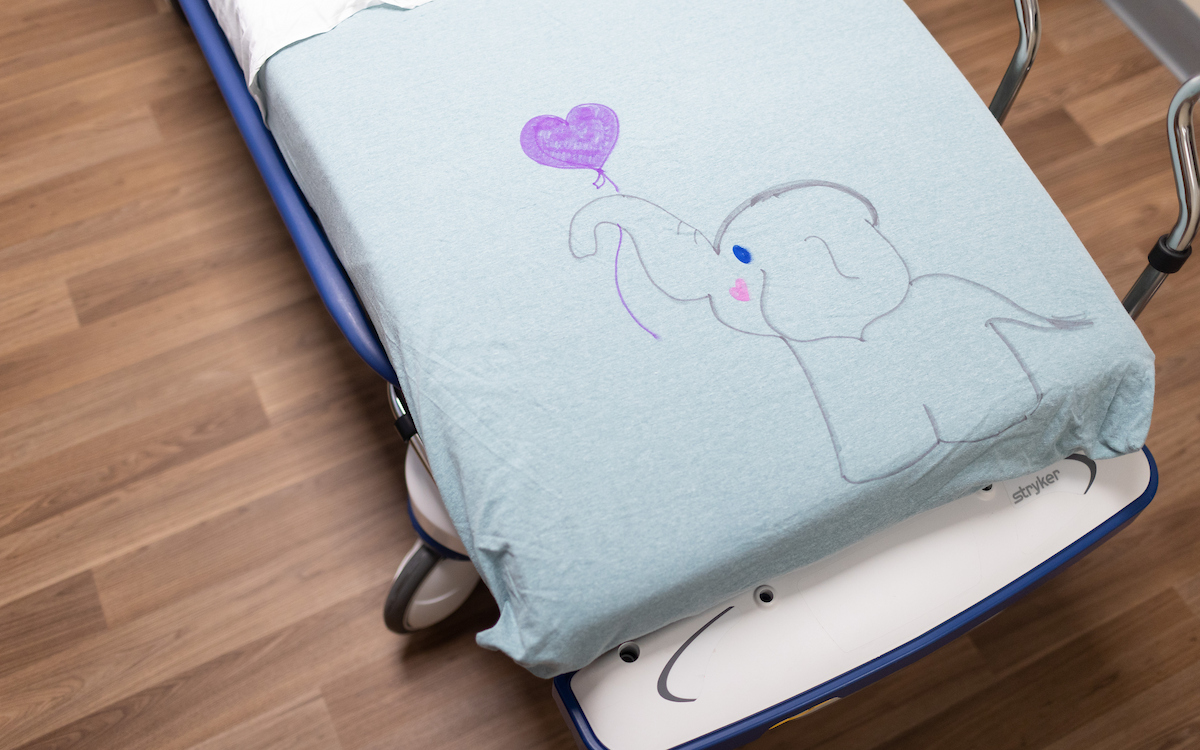 A light blue hospital sheet has a child's drawing of an elephant holding a purple heart balloon.

