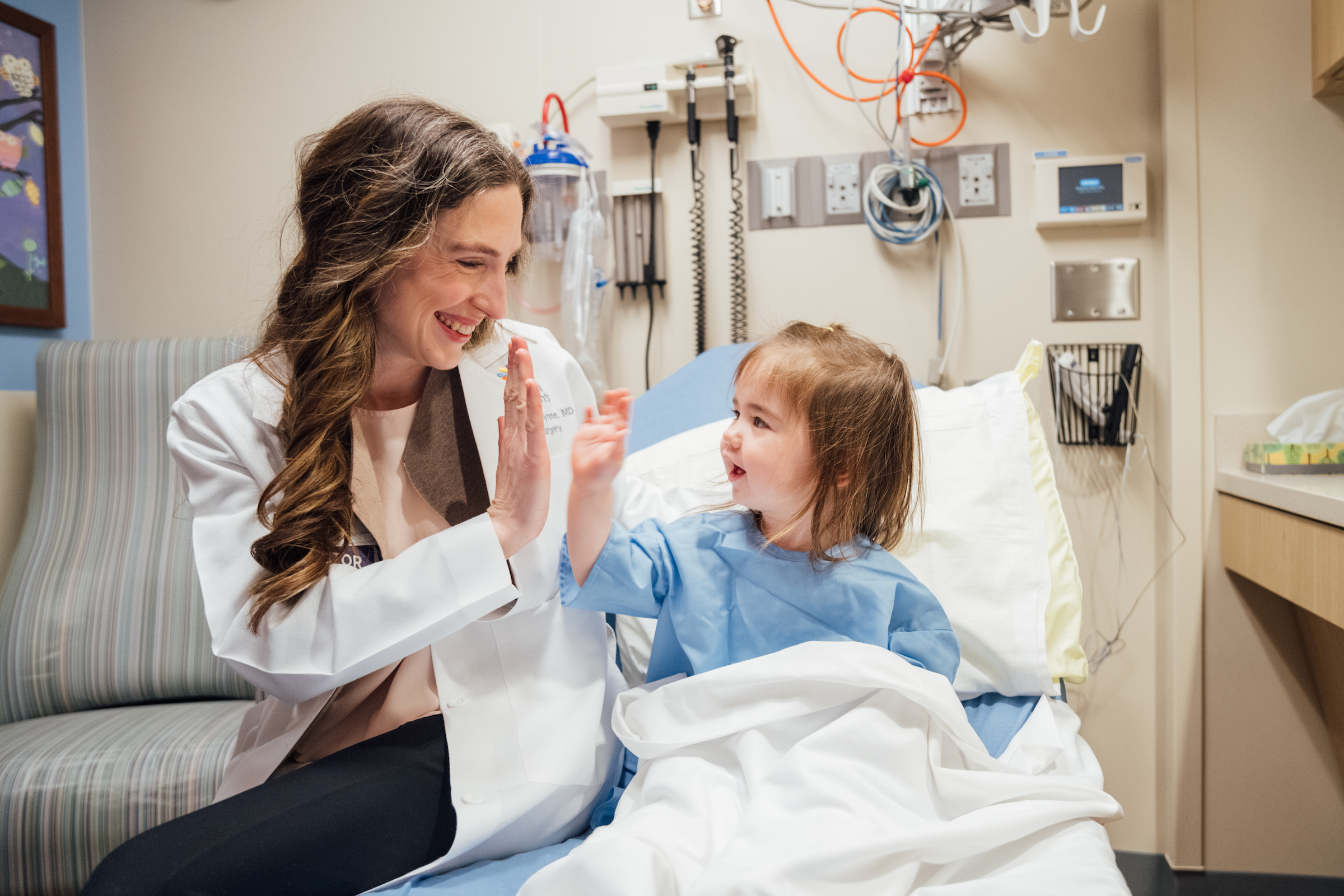 A female physician in a white coat gives a high-five to a smiling young girl in a blue hospital gown.
