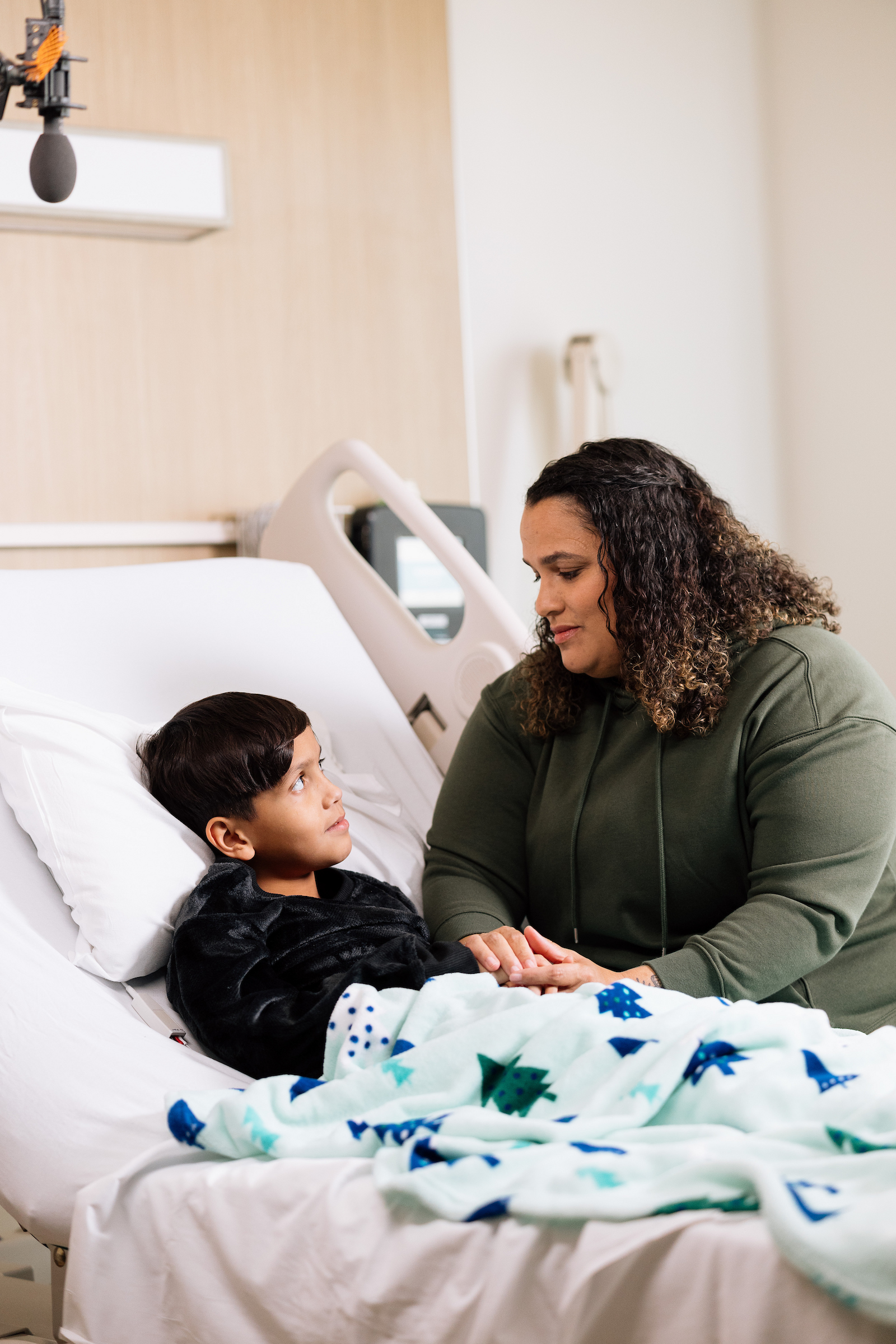 Parent sitting beside their child in a hospital bed, holding hands and offering comfort during recovery at a children’s hospital.

