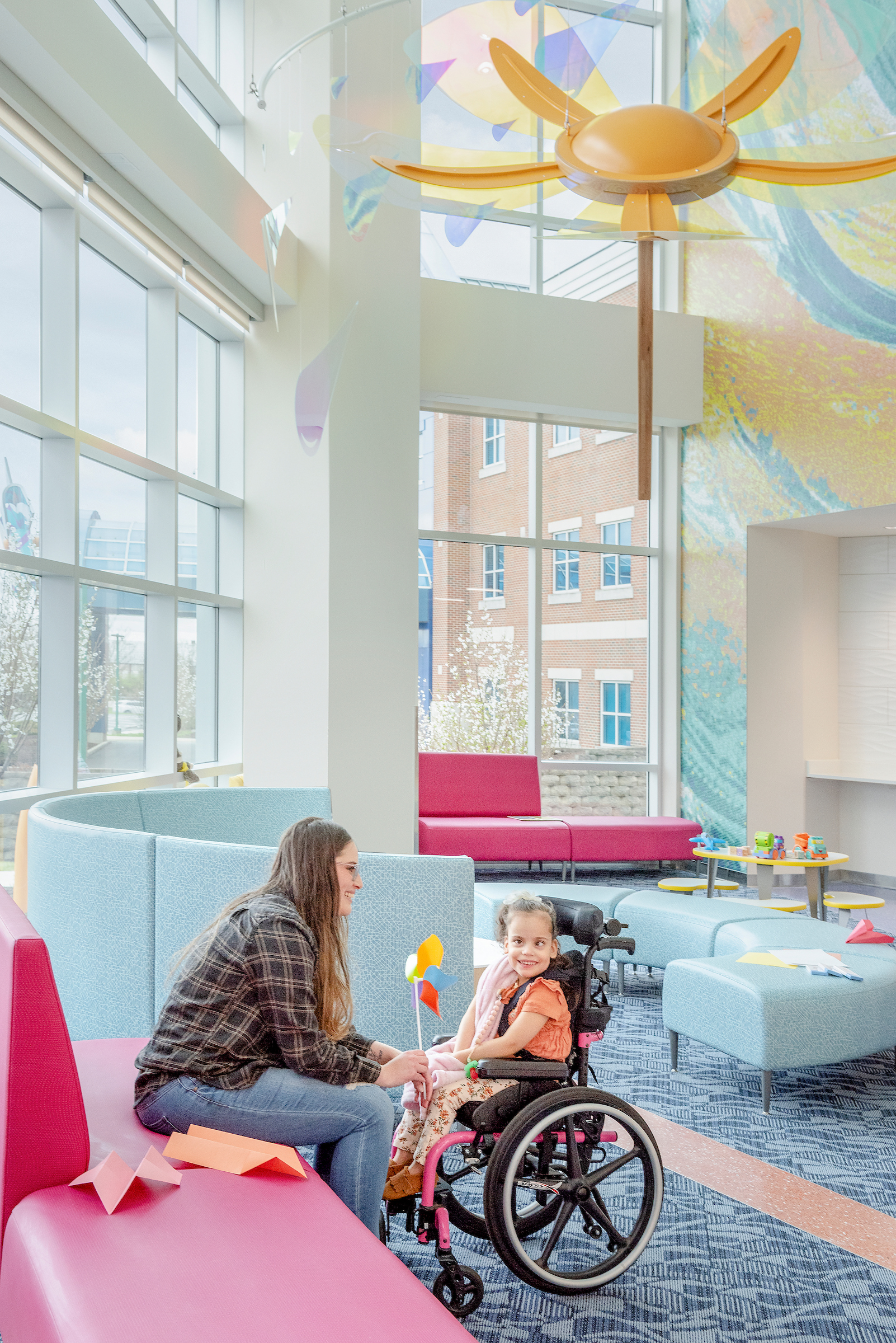 Parent sitting beside their child in a wheelchair in a bright hospital lobby, sharing a smile in the children’s rehabilitation center.