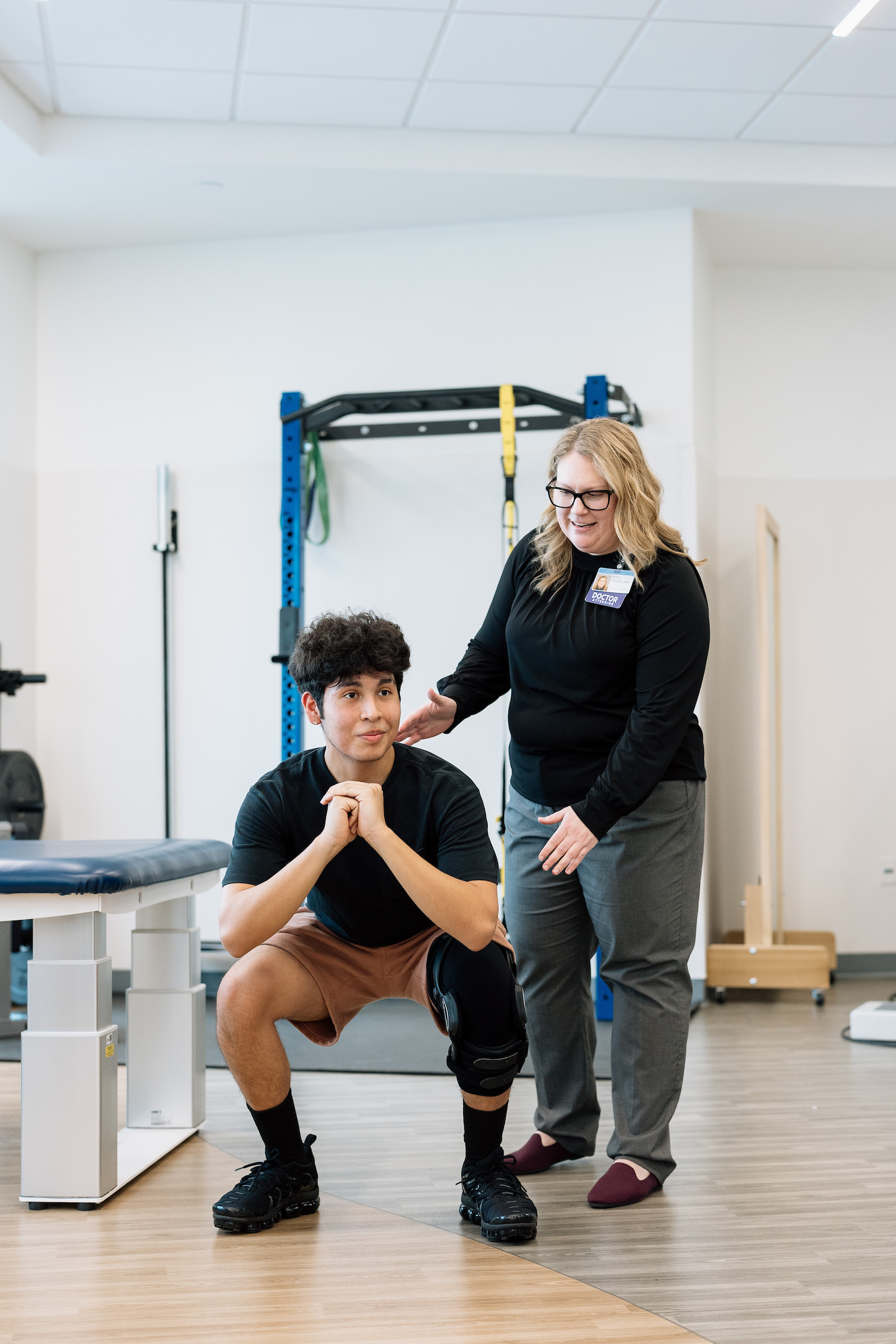 Physical therapist guiding a teenage patient through a squat exercise during a rehabilitation session in a physical therapy gym.

