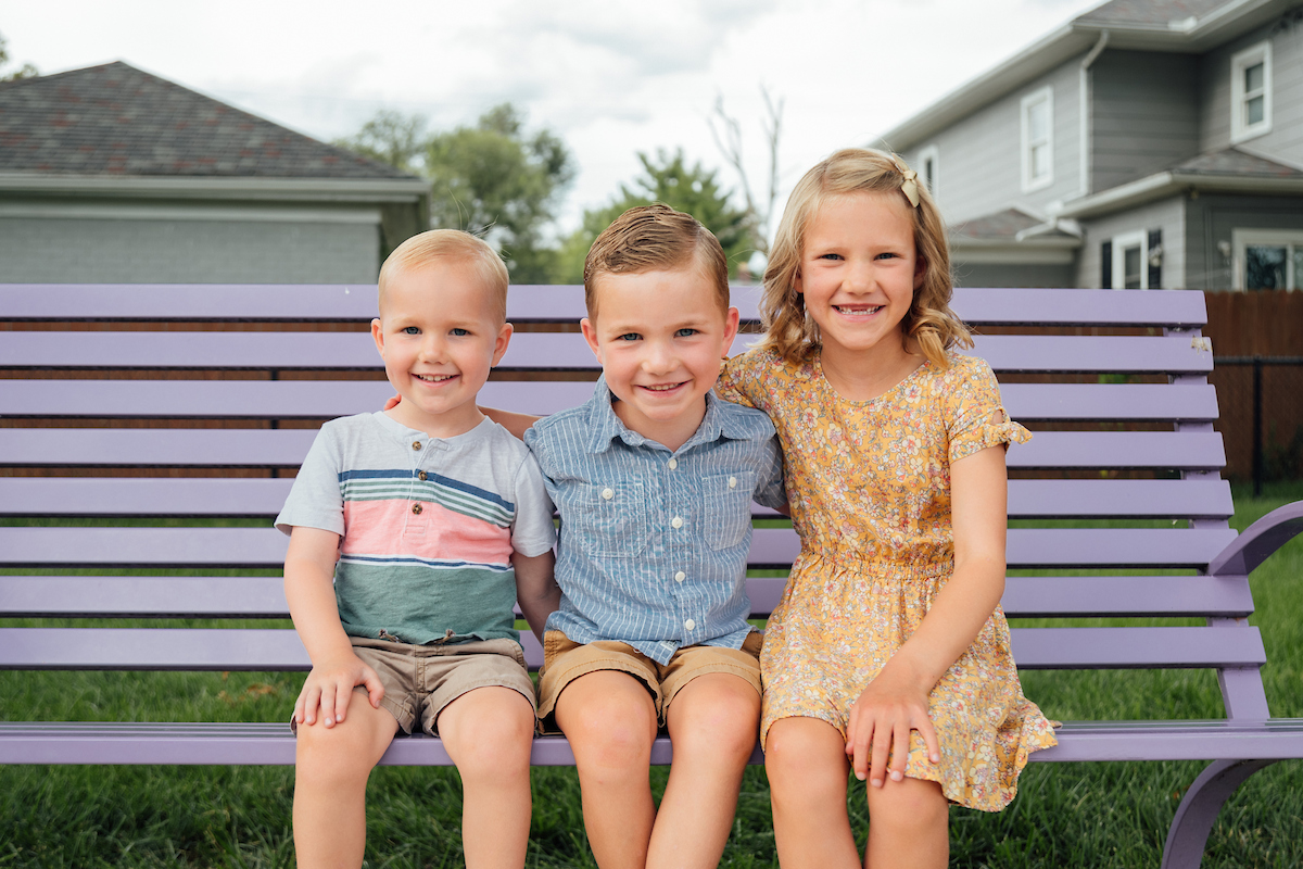  Three young children sit smiling together on a purple bench outdoors.


