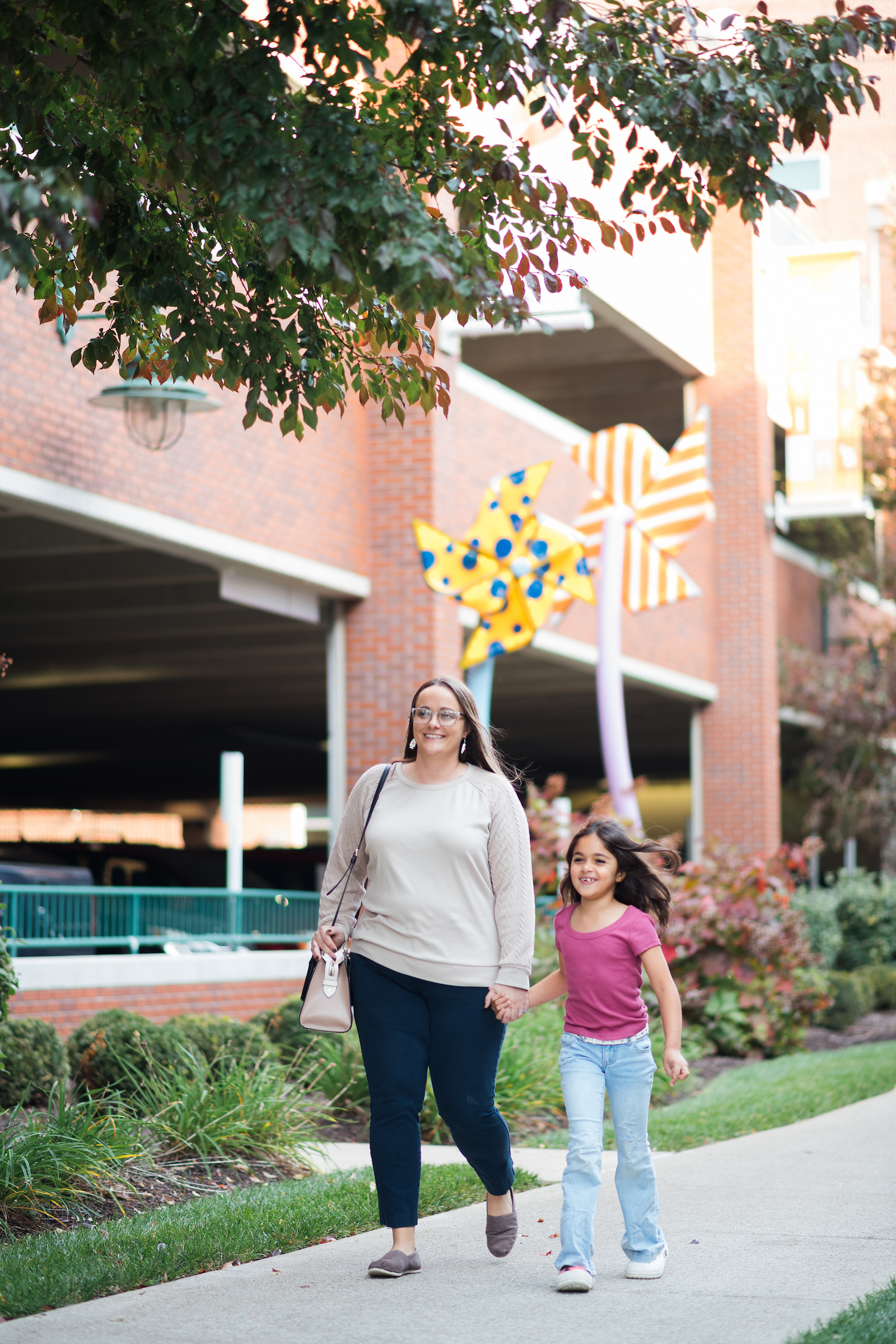 A smiling woman in glasses holds hands with a young girl as they walk past a brick building with large pinwheel sculptures.
