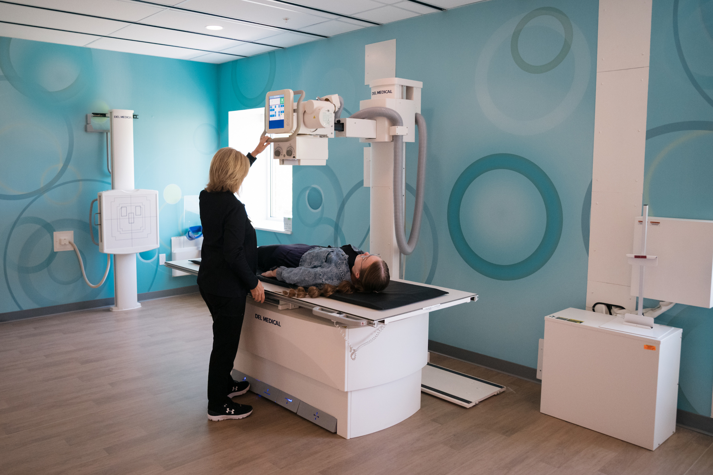 A medical professional adjusts an X-ray machine over a patient lying on a table in a room with blue patterned walls.

