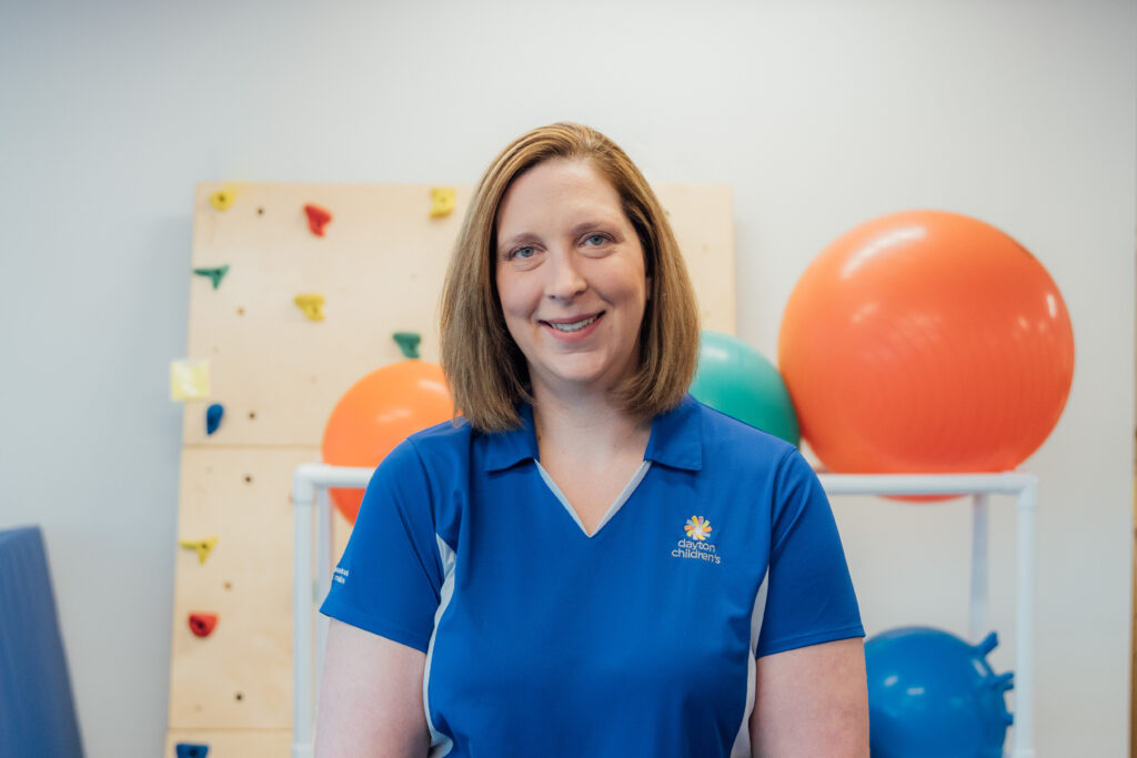  A friendly female healthcare professional with medium-length hair smiles in a blue polo shirt in a therapy gym, with exercise equipment behind her.