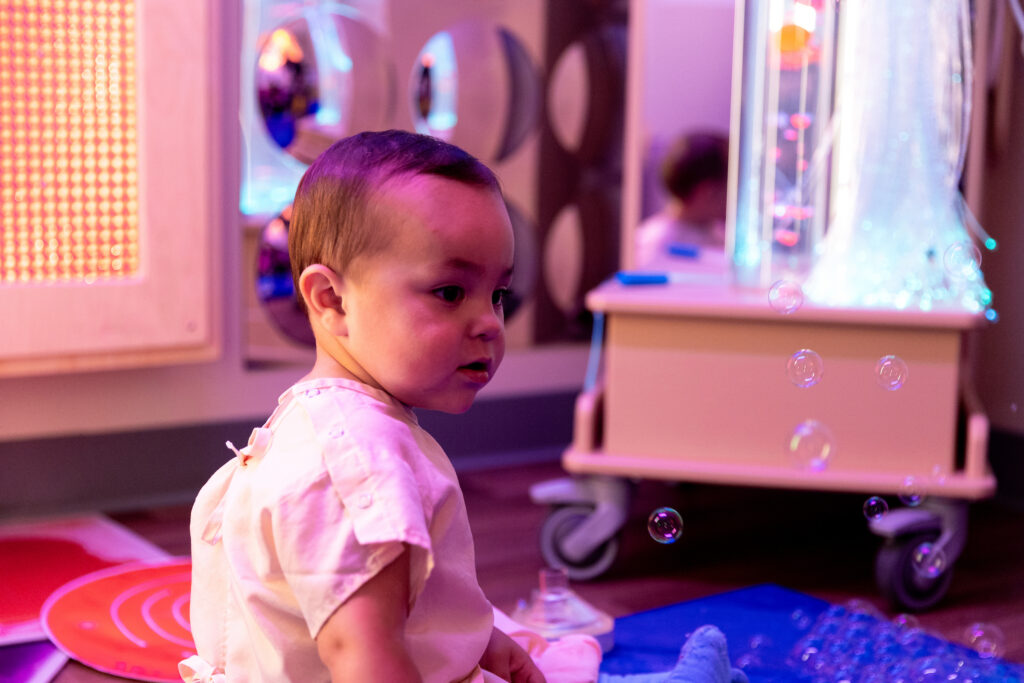 A baby in a light-colored shirt looks at floating bubbles in a softly lit sensory room with a bubble tube.
