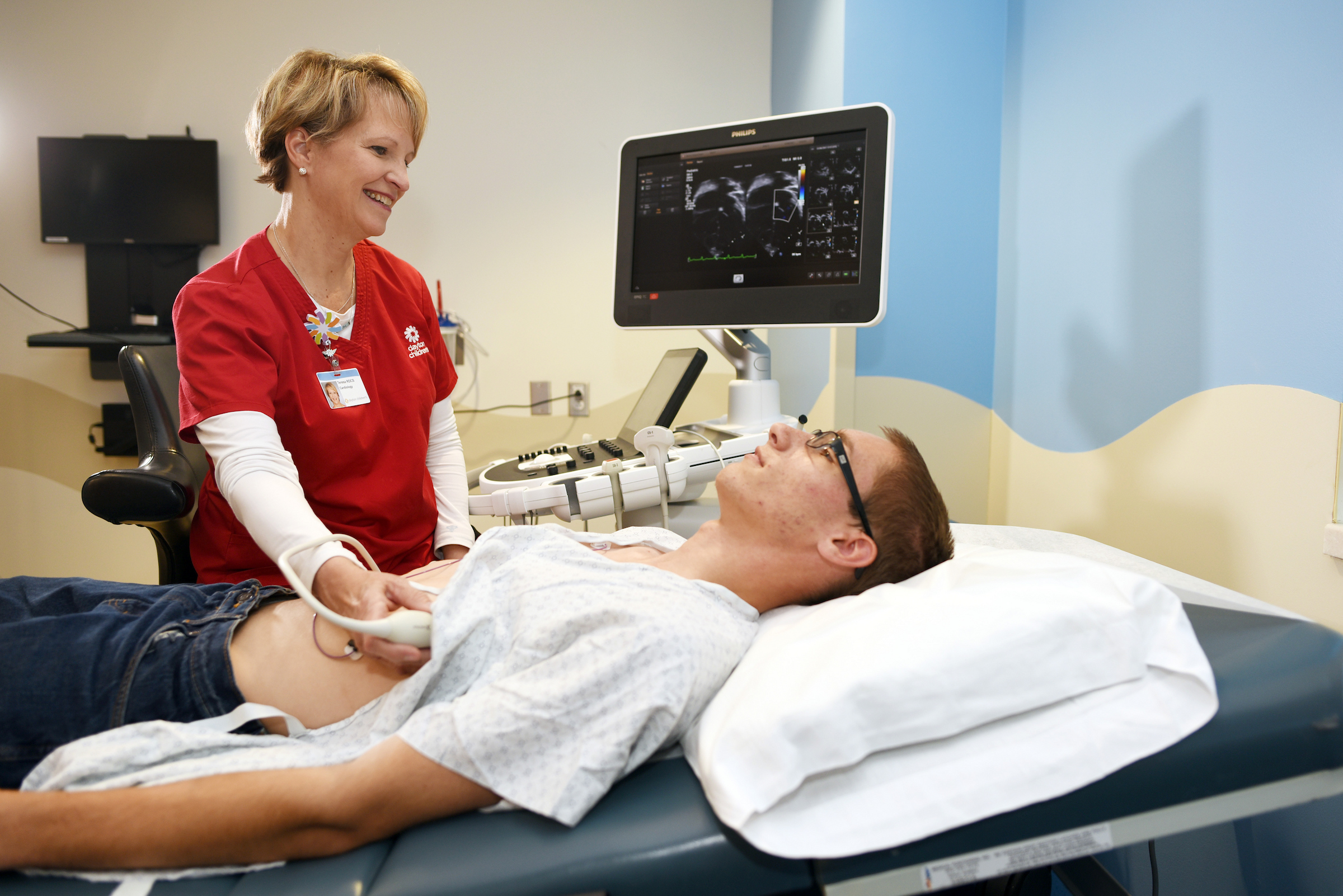 A smiling female medical professional performs an ultrasound on a male patient lying on an exam table.
