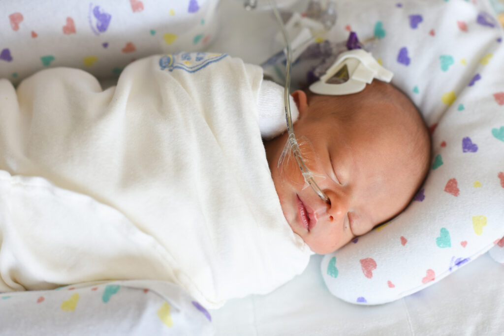 A newborn baby sleeps peacefully, swaddled in white, with a breathing tube and monitors visible.

