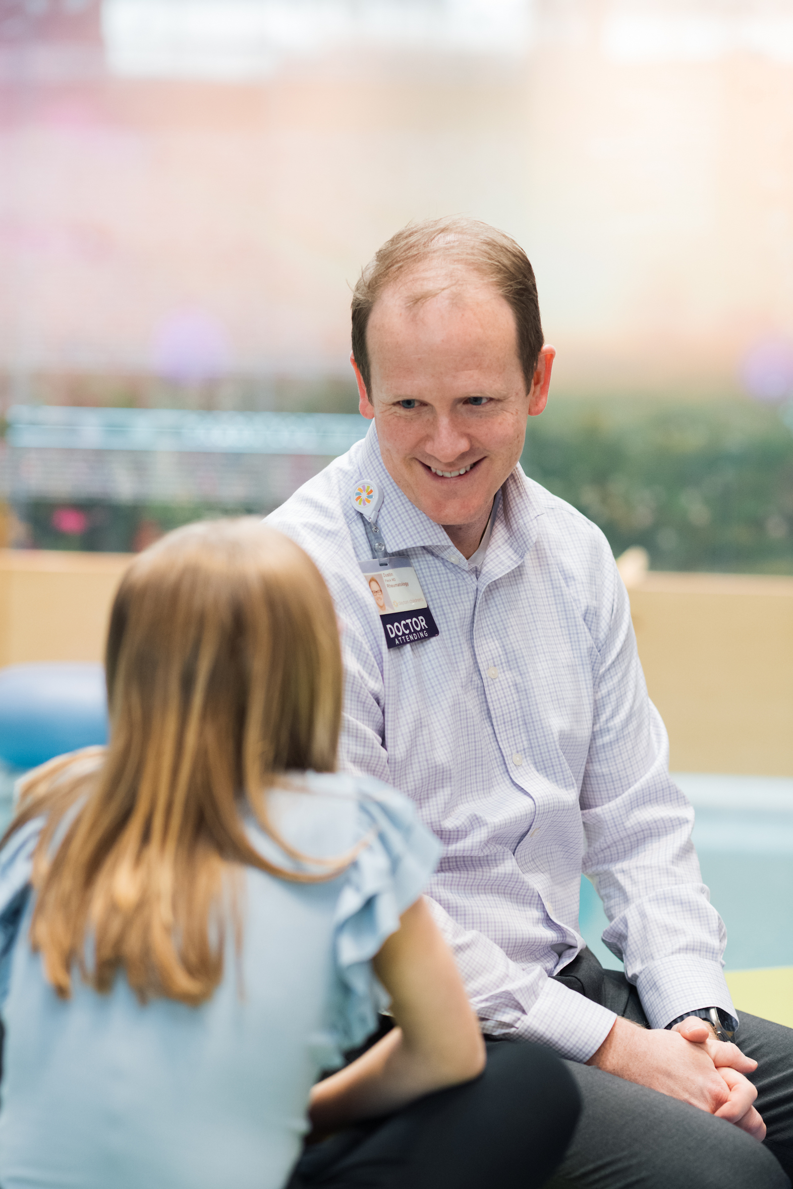 A smiling male doctor in a plaid shirt and "Doctor Attending" badge talks to a young patient, whose back is to the camera.