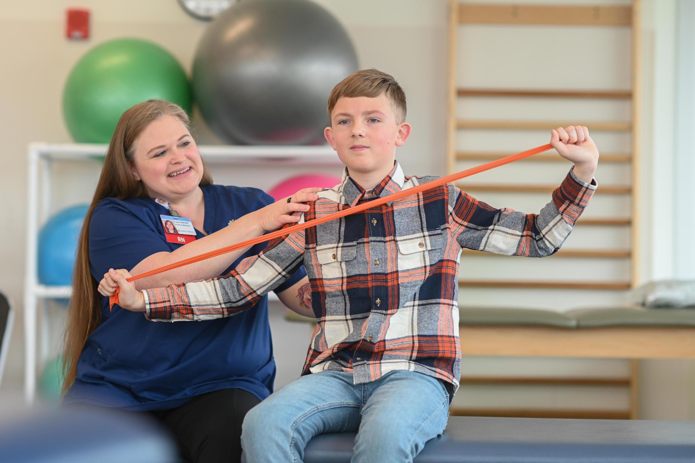 Nurse assisting a young boy with a resistance band exercise during a physical therapy session in a pediatric rehabilitation gym.
