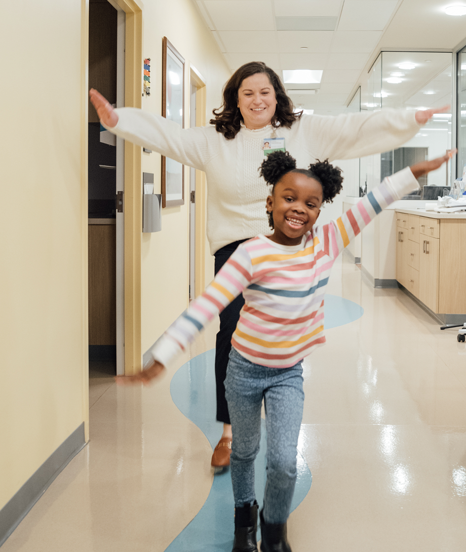 Child and caregiver joyfully walking down a hospital hallway, arms outstretched and smiling.