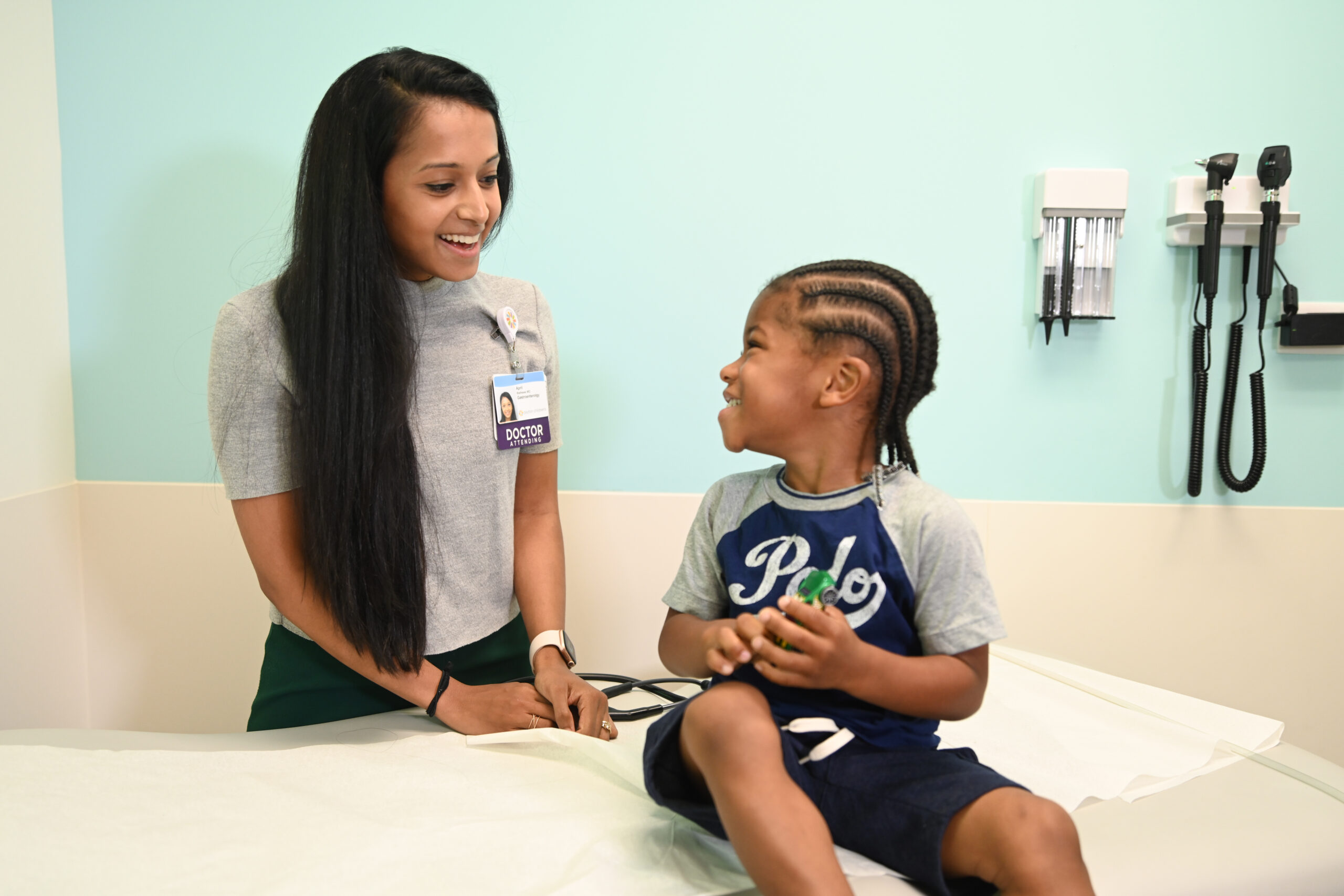 young boy sitting on patient bed smiling and looking back at female pediatric gastroenterologist 