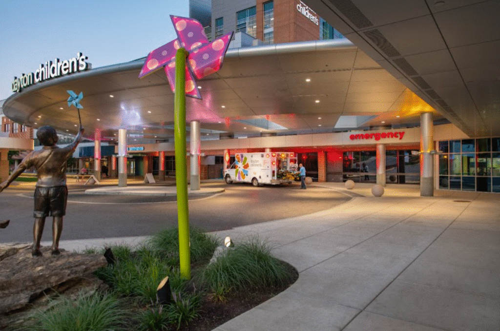 Exterior view of Dayton Children's Hospital emergency entrance at dusk, with an ambulance, a child statue, and a colorful pinwheel sculpture.