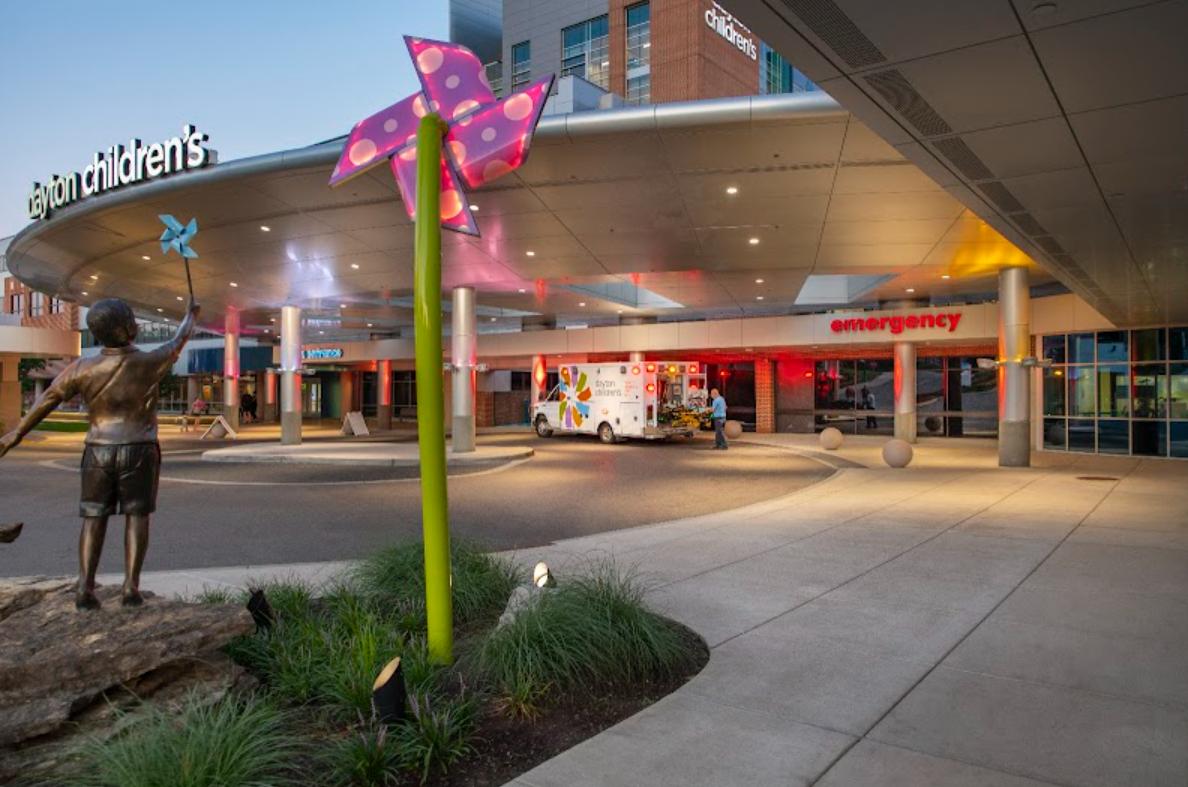Exterior view of Dayton Children's Hospital emergency entrance at dusk, with an ambulance, a child statue, and a colorful pinwheel sculpture.