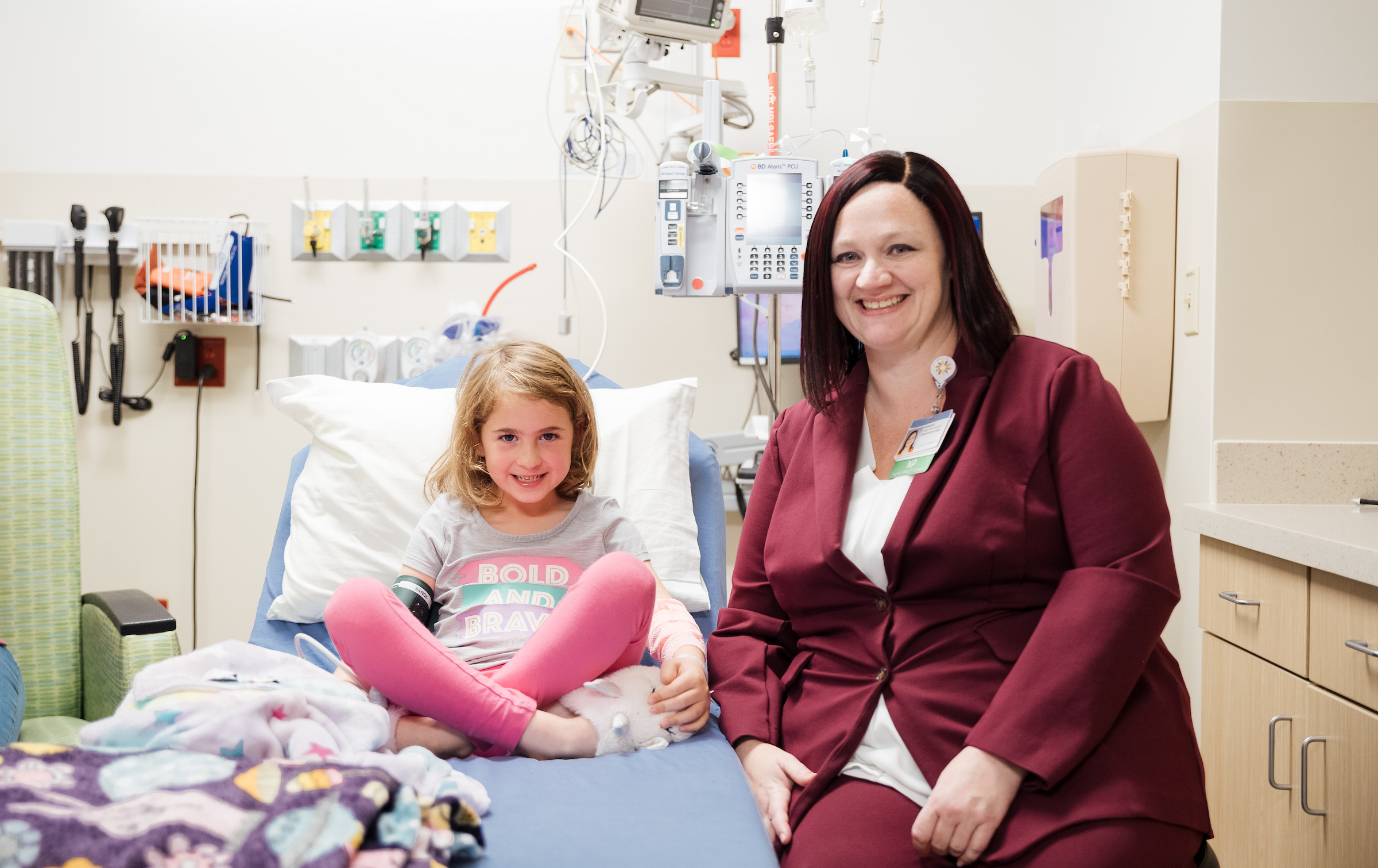 A concerned mother talks to her young daughter, who looks down with a hand on her stomach possibly due to discomfort from inflammatory bowel disease. 
