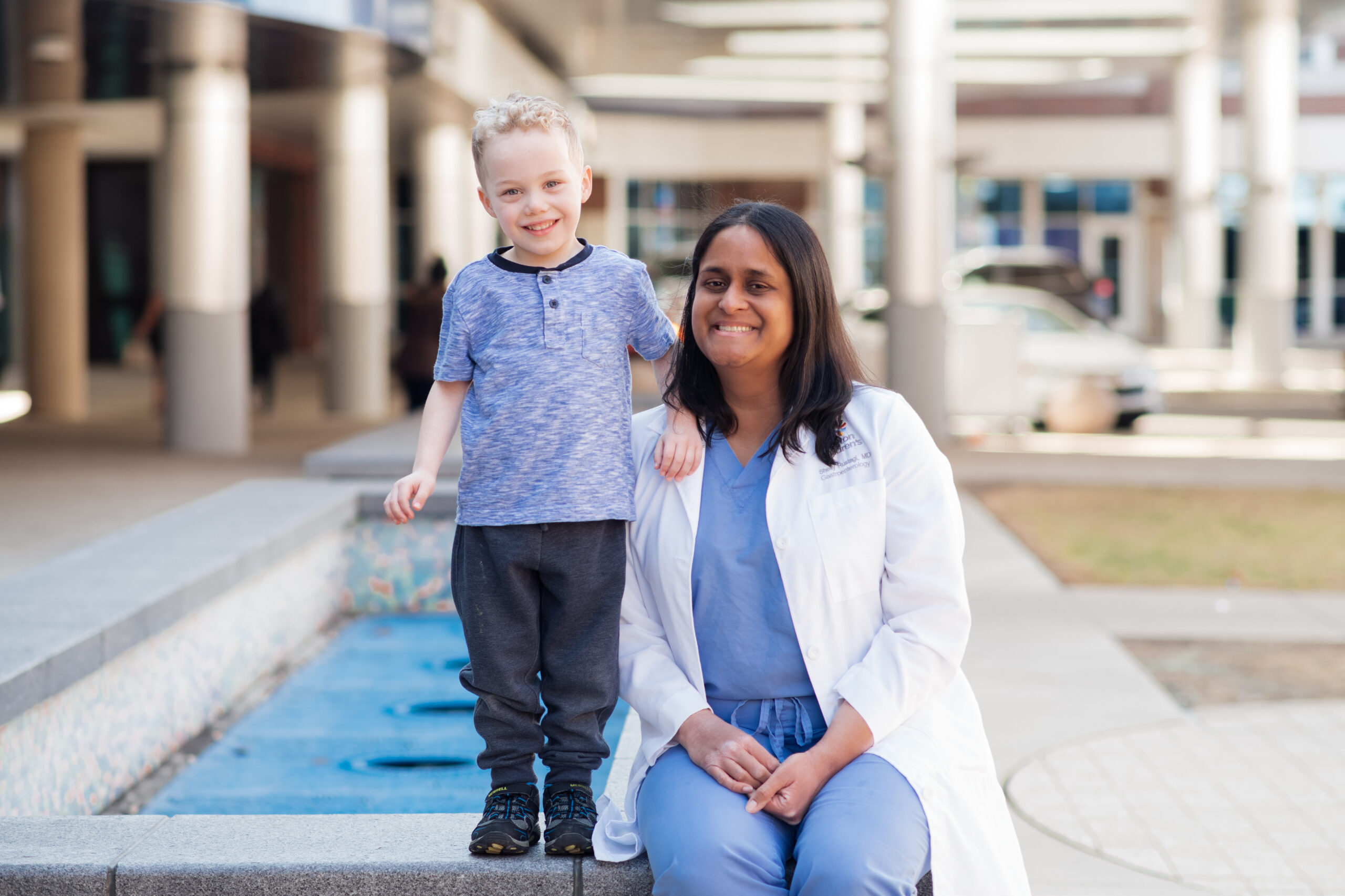 Dr. Shelly Rustagi, Pediatric Gastroenterologist, sitting by fountain with boy patient next to her smiling 