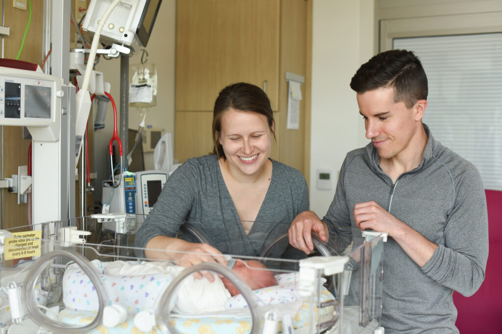 A happy mom and dad look down at their baby in its bed in the NICU.