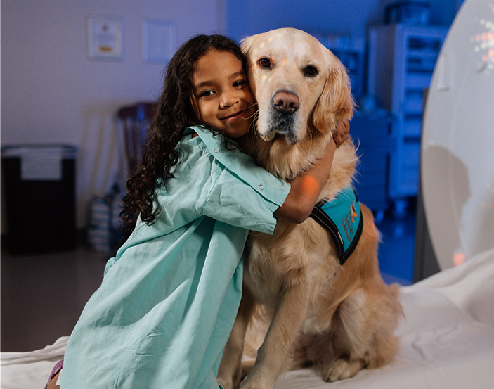  A young girl in a hospital gown hugs a golden retriever therapy dog, showing the comfort and emotional support animals provide in pediatric care settings.