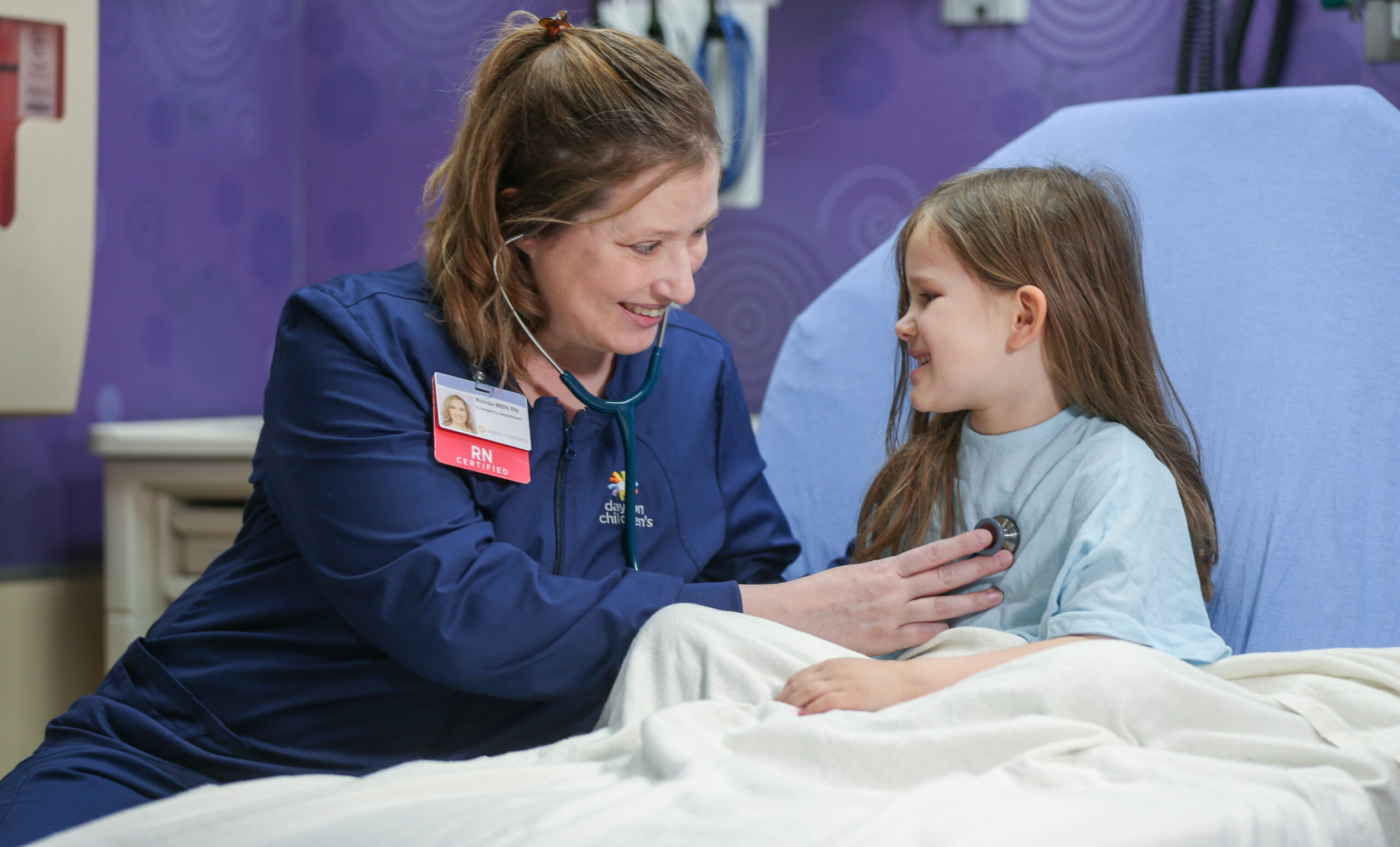 A smiling nurse listens to a young girl's heart with a stethoscope in a hospital room