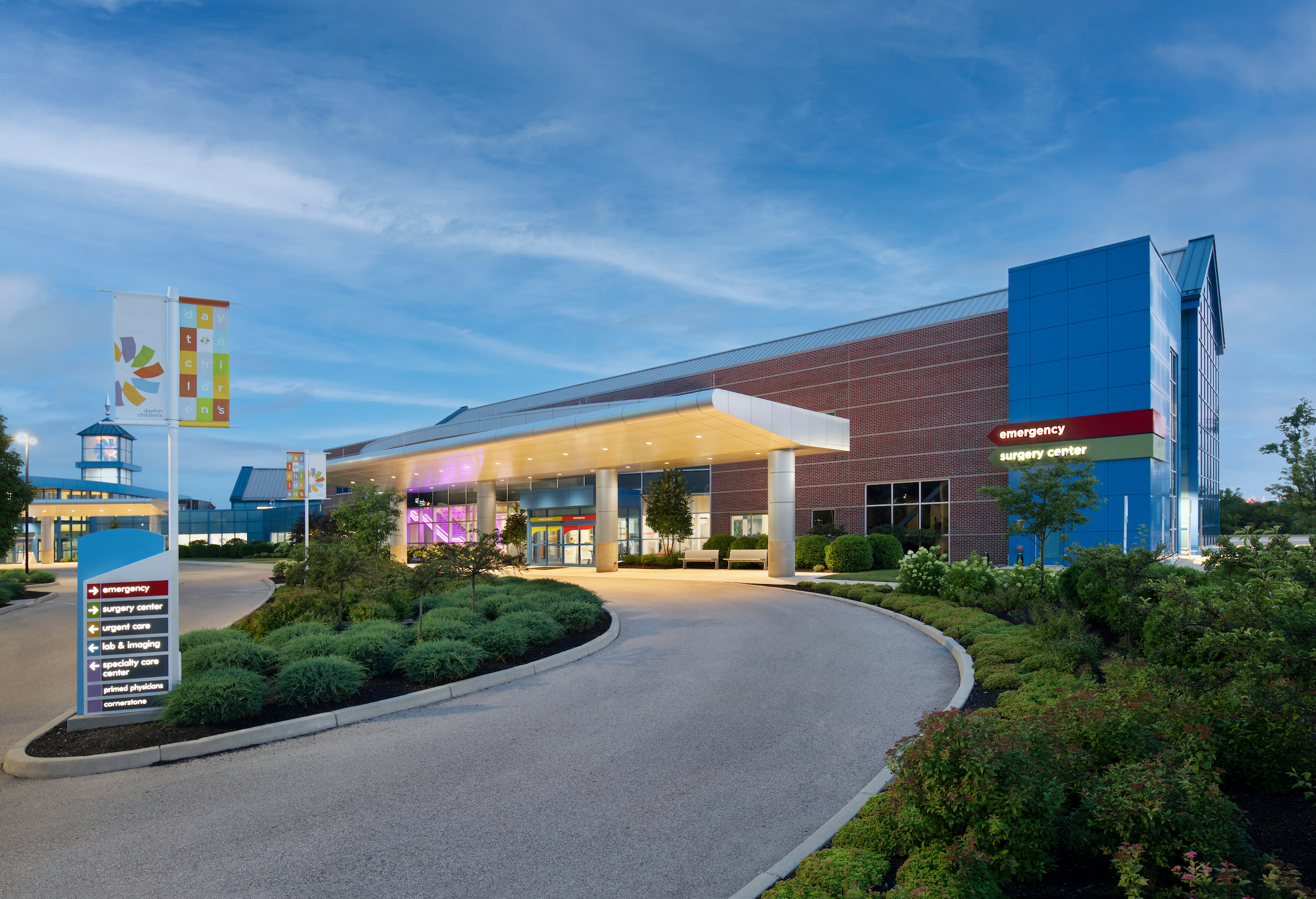 Exterior of a modern hospital entrance with 'emergency' and 'surgery center' signs, under a twilight sky.
