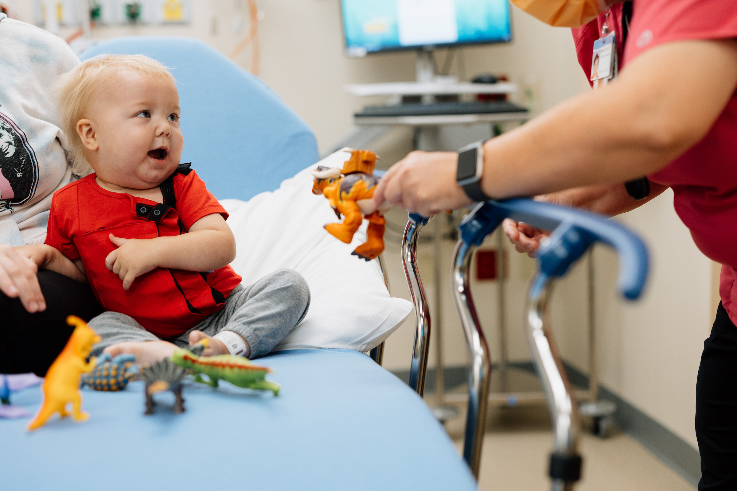 A baby on a hospital bed looks surprised as an adult in red scrubs offers a toy dinosaur