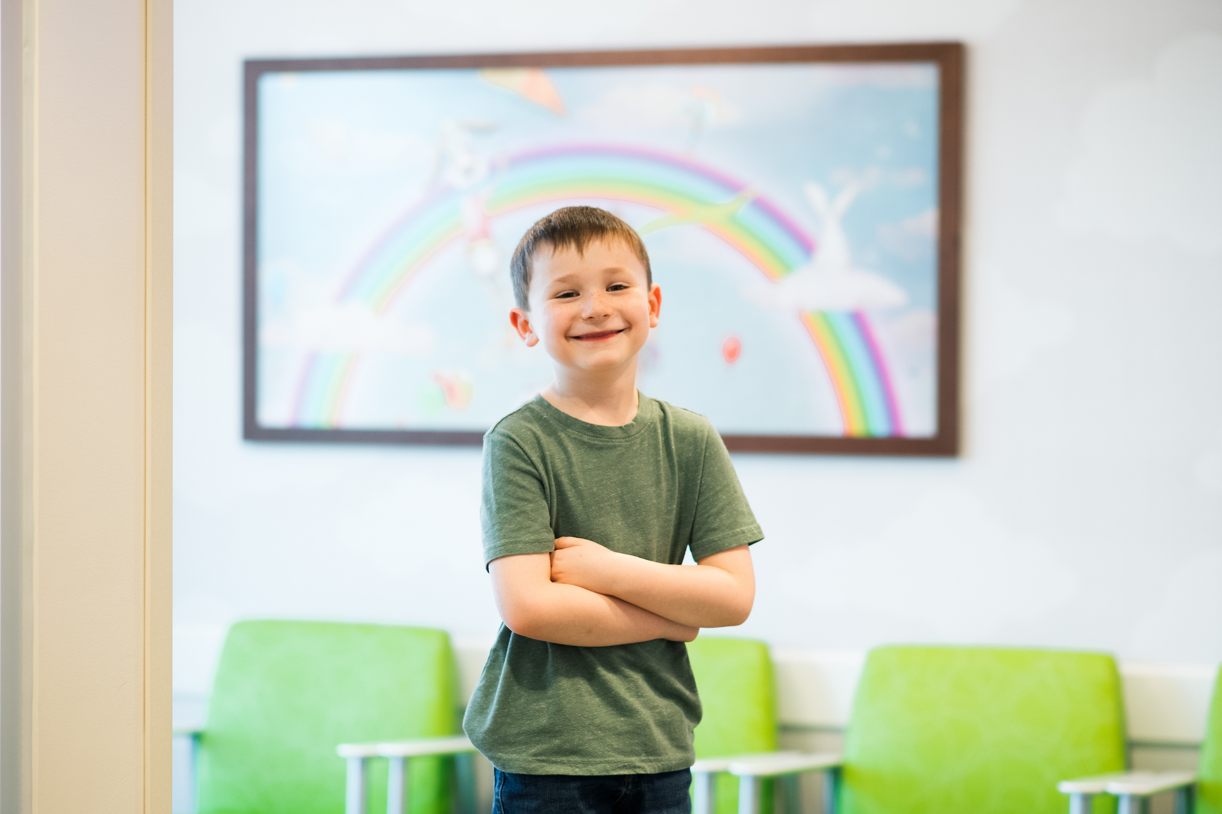 A young boy in a green t-shirt smiles with his arms crossed in front of a colorful rainbow poster.