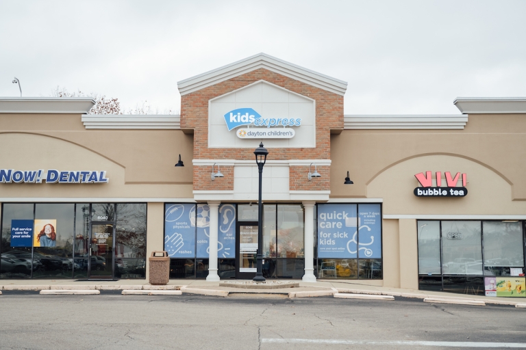 Exterior of a 'Kids Express by Dayton Children's' urgent care clinic, flanked by other storefronts, under a cloudy sky.