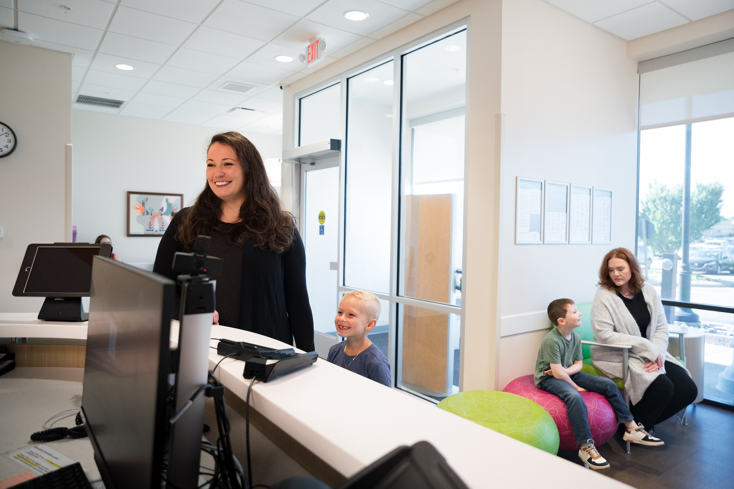 A smiling receptionist at a medical front desk, with children and an adult waiting in the background.