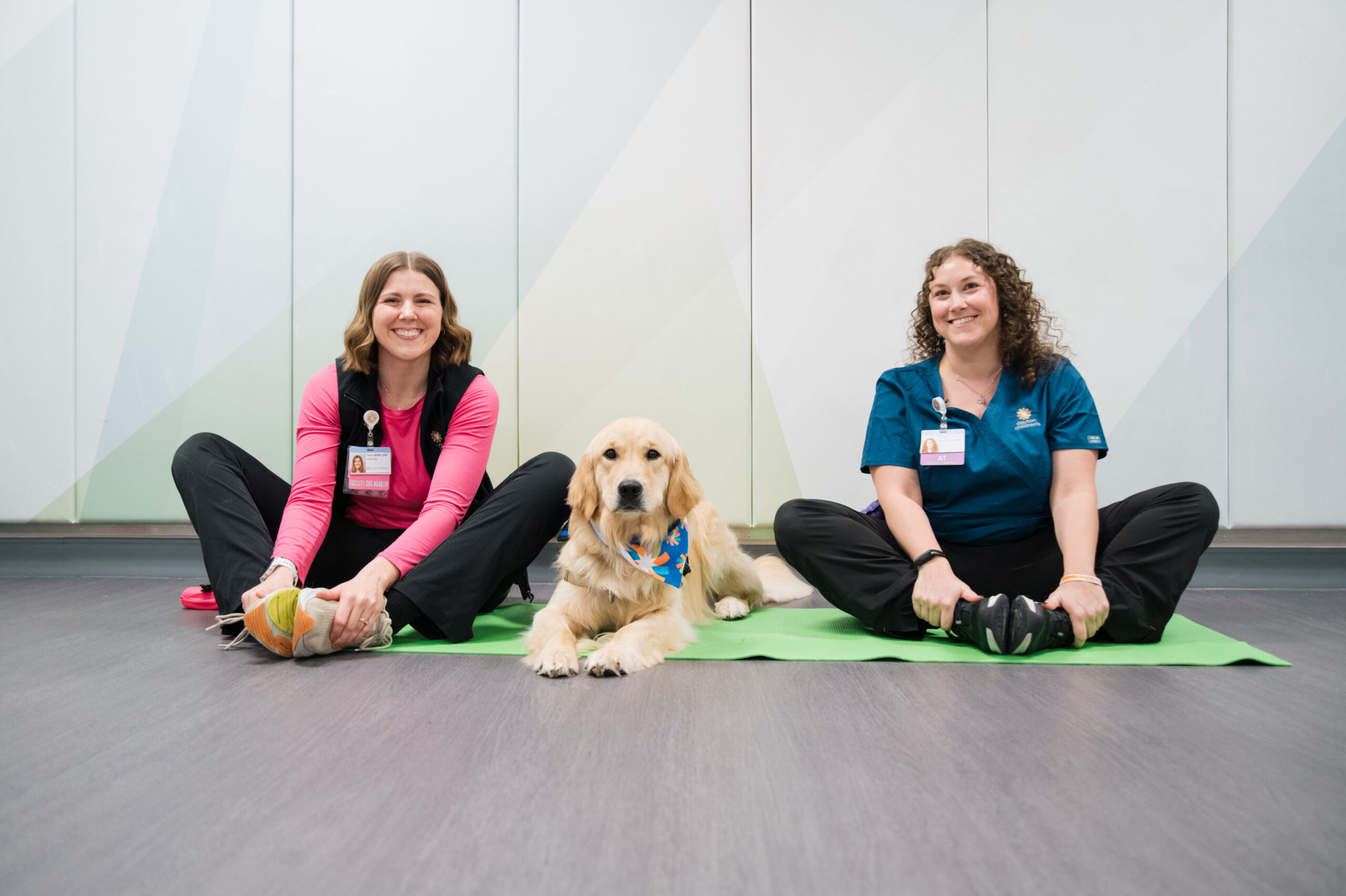 A golden retriever lies between two smiling women in athletic wear, sitting cross-legged on a green mat.


