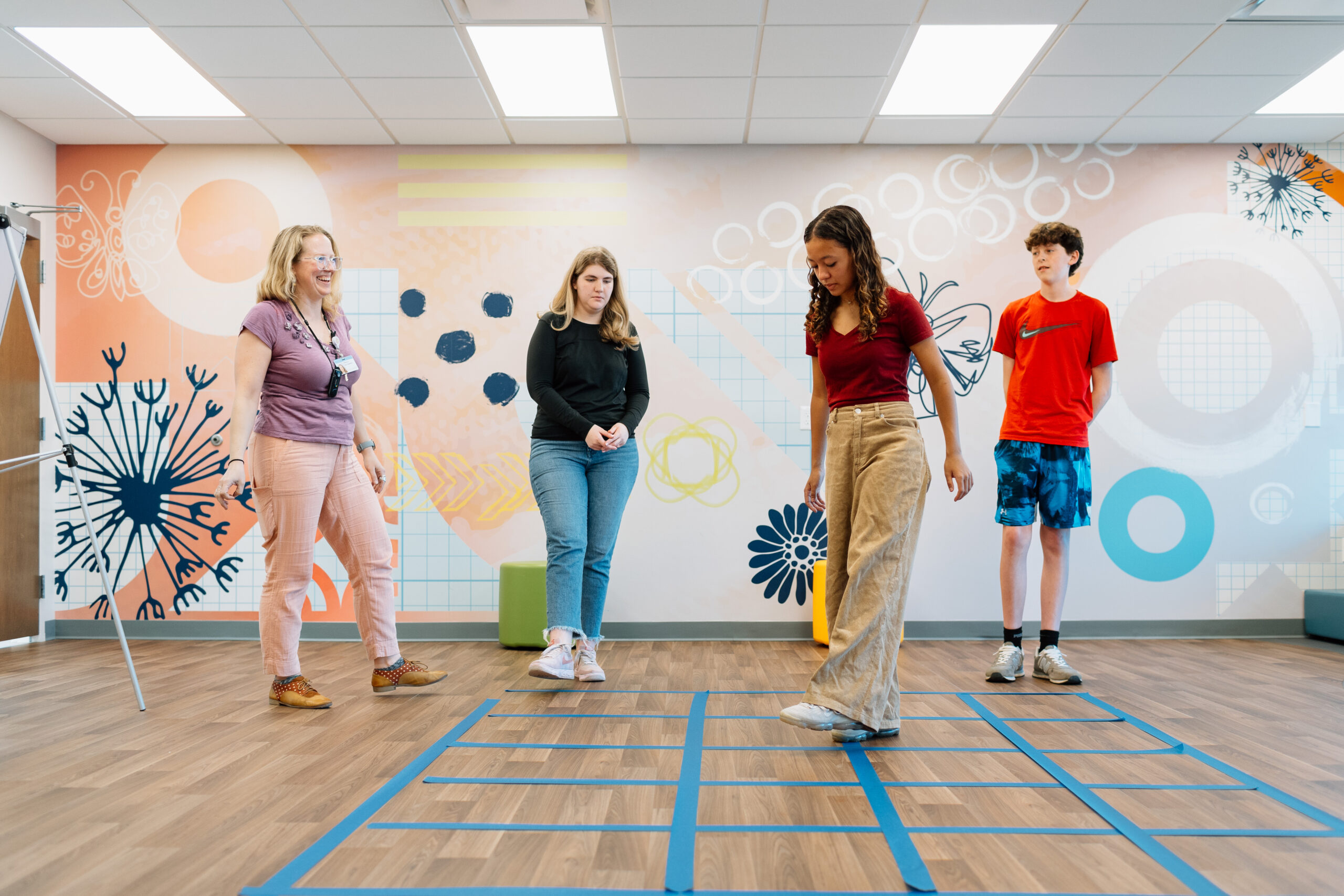 An adult and three teenagers interact with a grid marked on the floor, in a brightly decorated room.

