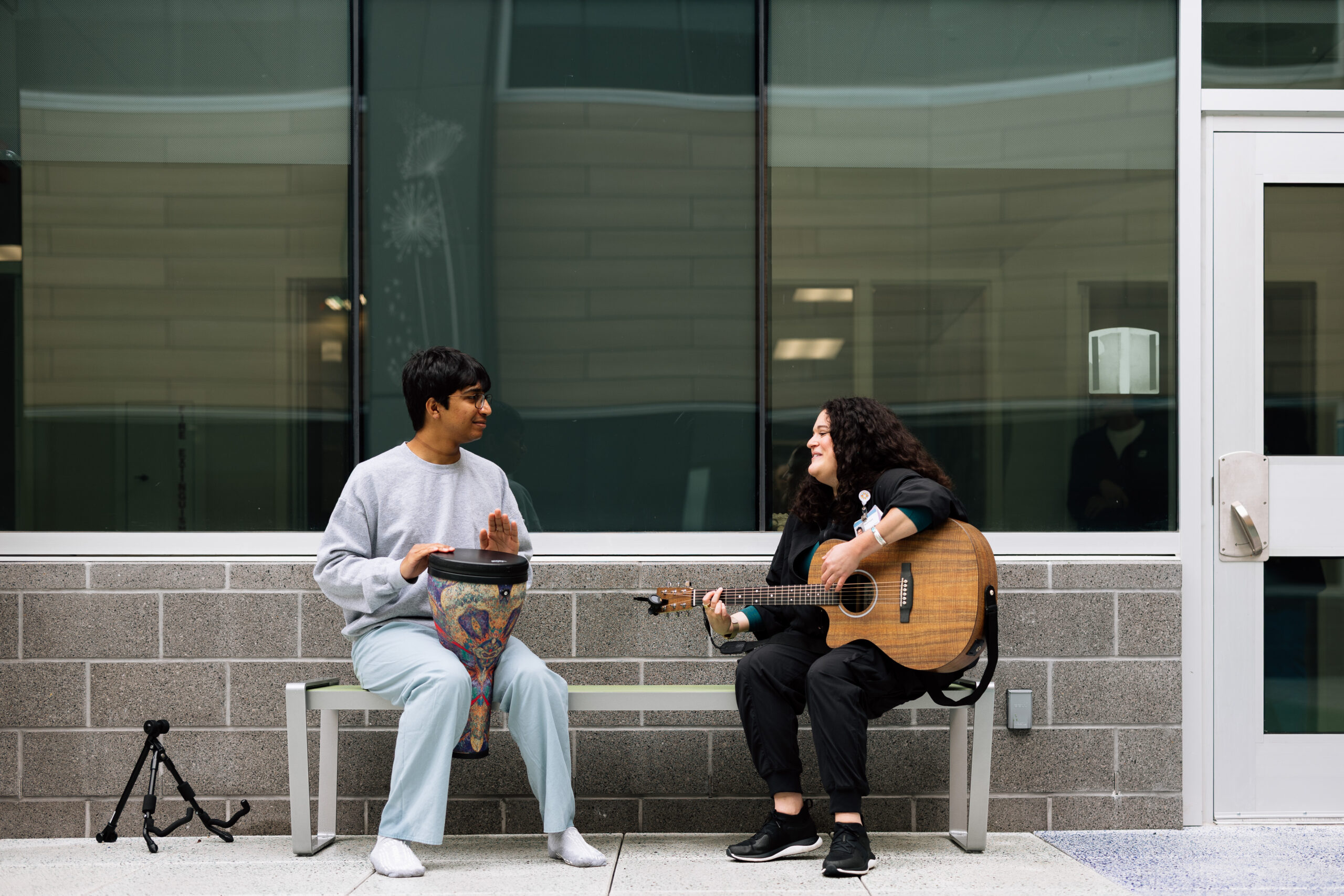 Two people sit on a bench outside a building; one plays a djembe drum, and the other plays an acoustic guitar.