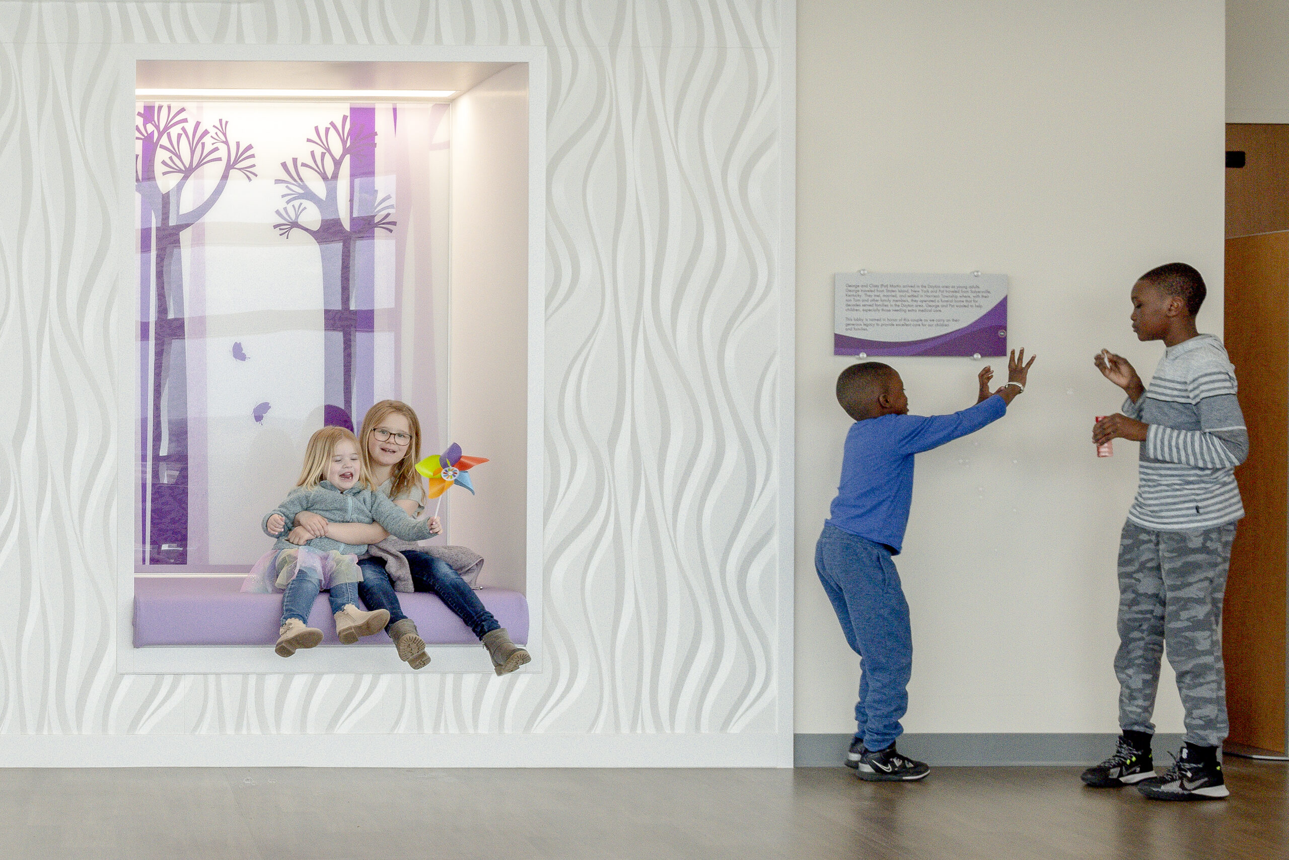 Two girls sit in a purple-lit alcove with a tree mural, while two boys play with bubbles nearby.

