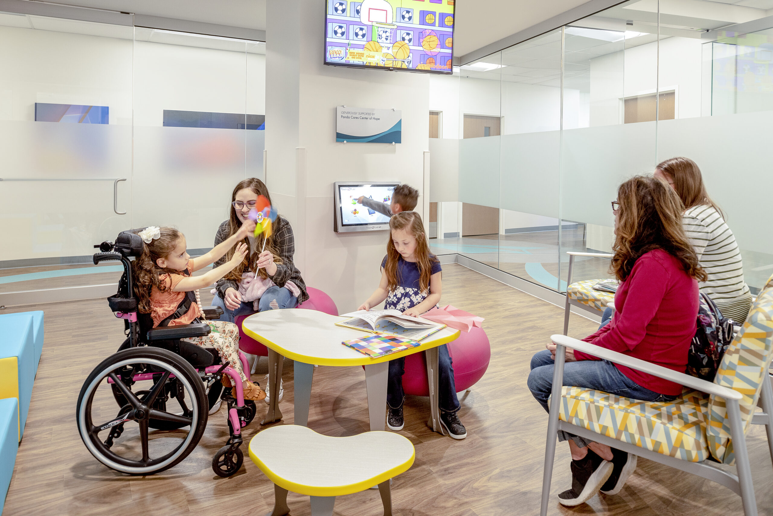 A girl in a wheelchair plays with a pinwheel while a woman holds another child, and another girl reads at a table in a waiting area.