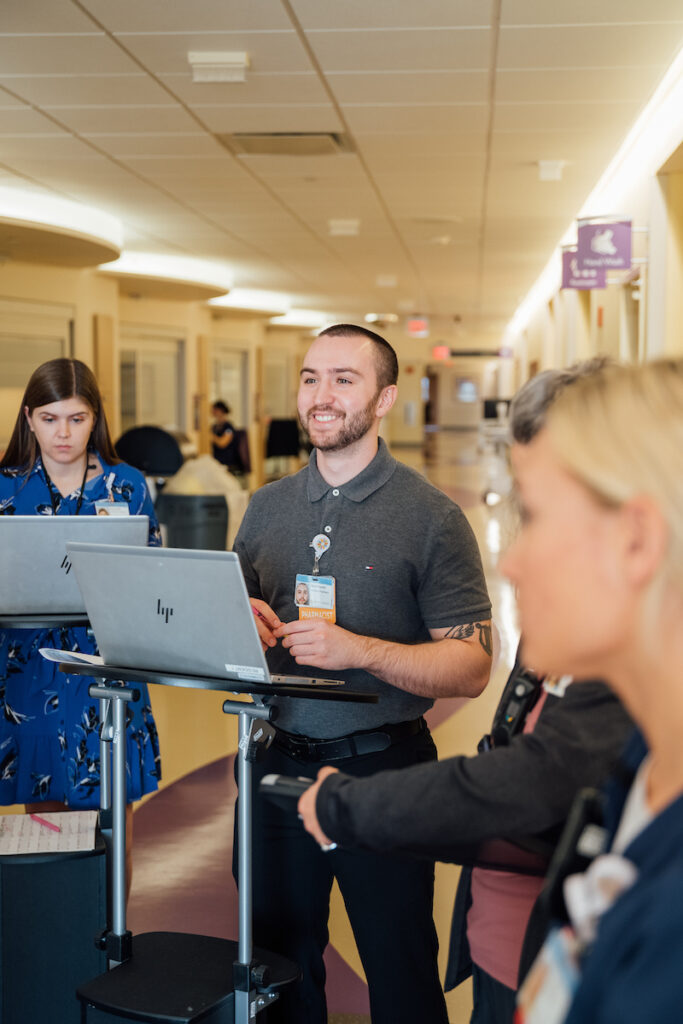 A smiling male medical professional in a gray polo shirt stands at a laptop cart in a hospital hallway with colleagues.

